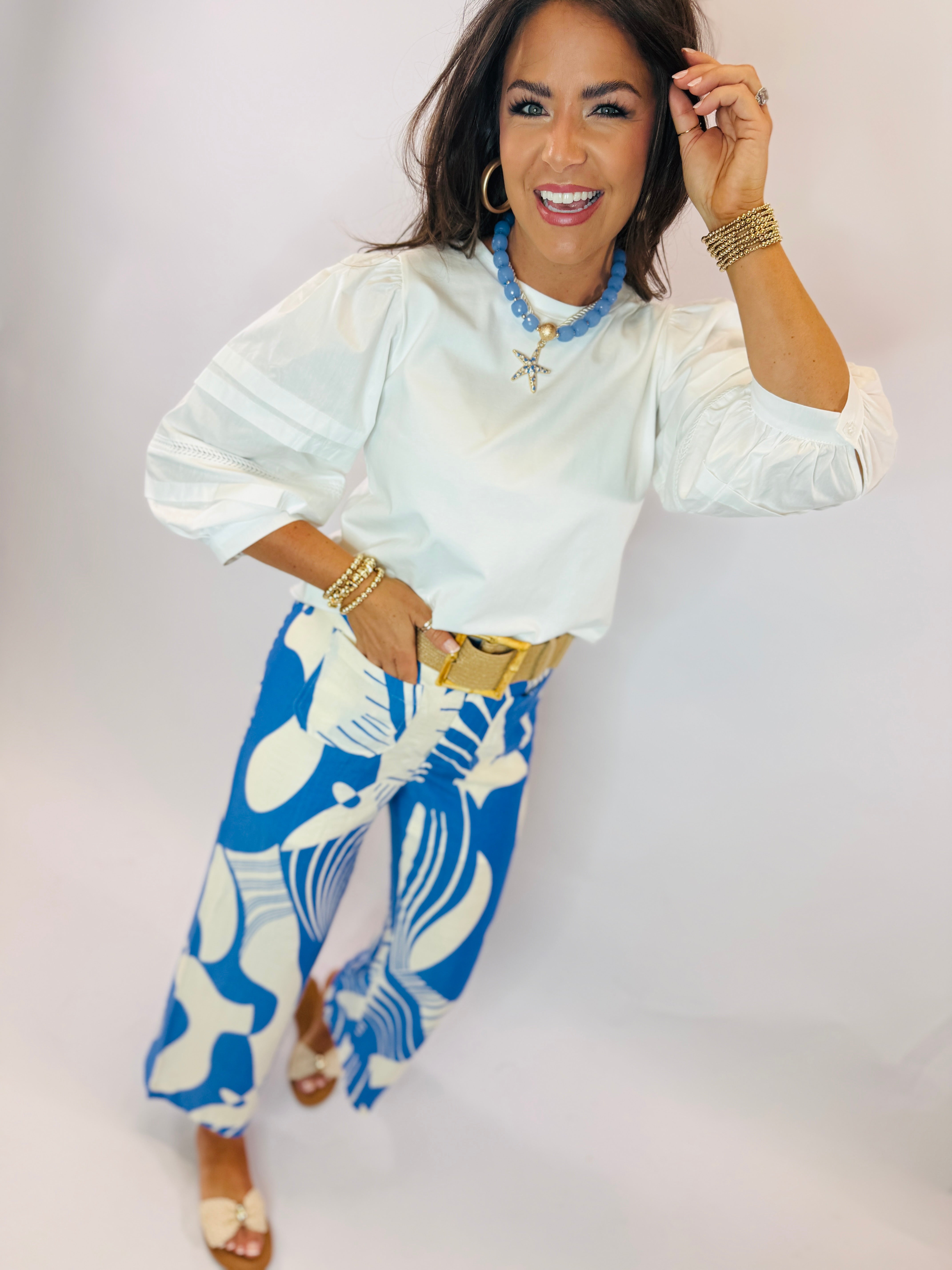 Woman wearing white Carol top with blue beaded necklace, paired with blue and white patterned skirt and gold accessories.