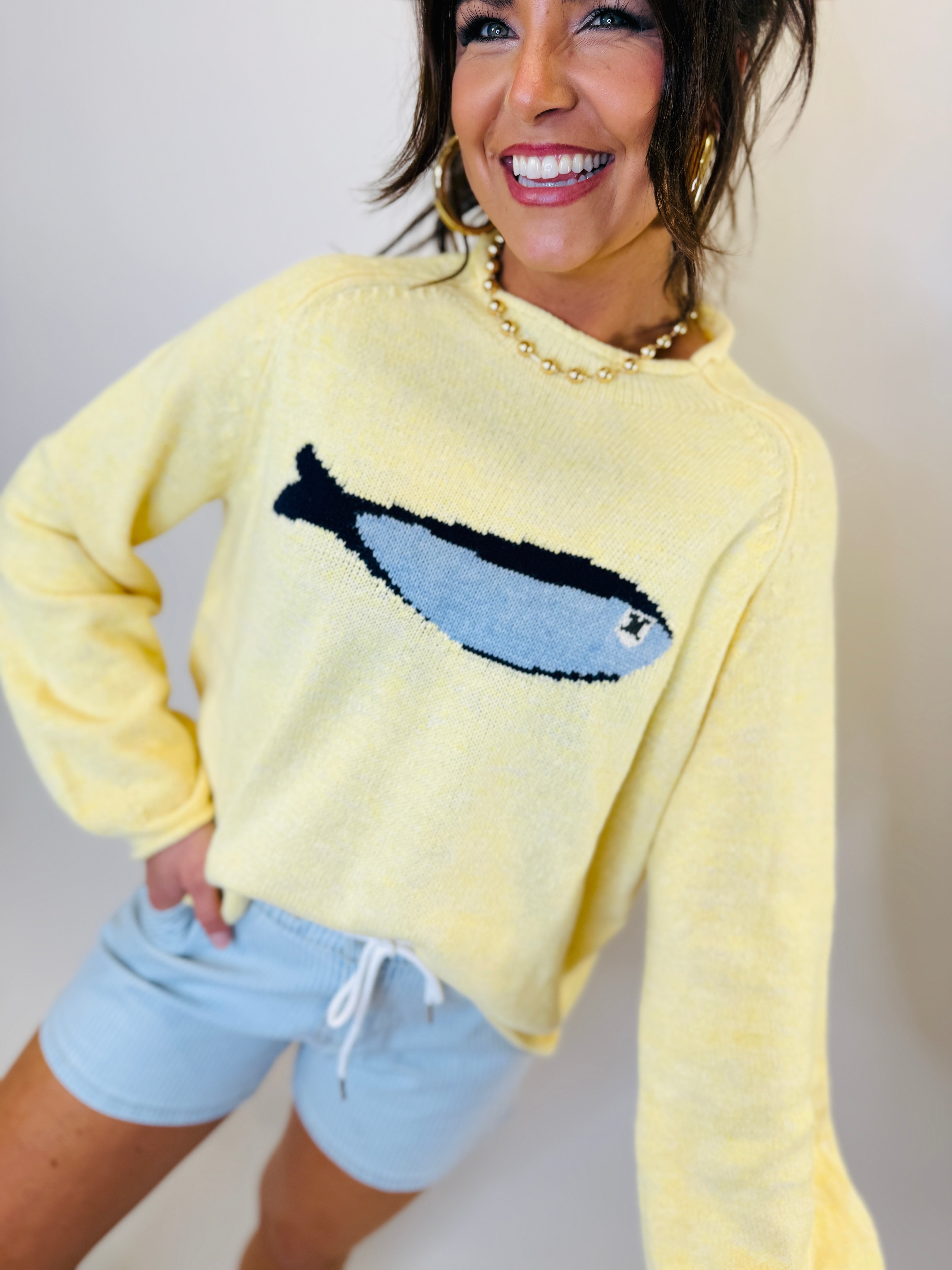 Woman wearing yellow knit sweater with blue whale design and light blue drawstring shorts, smiling at camera.