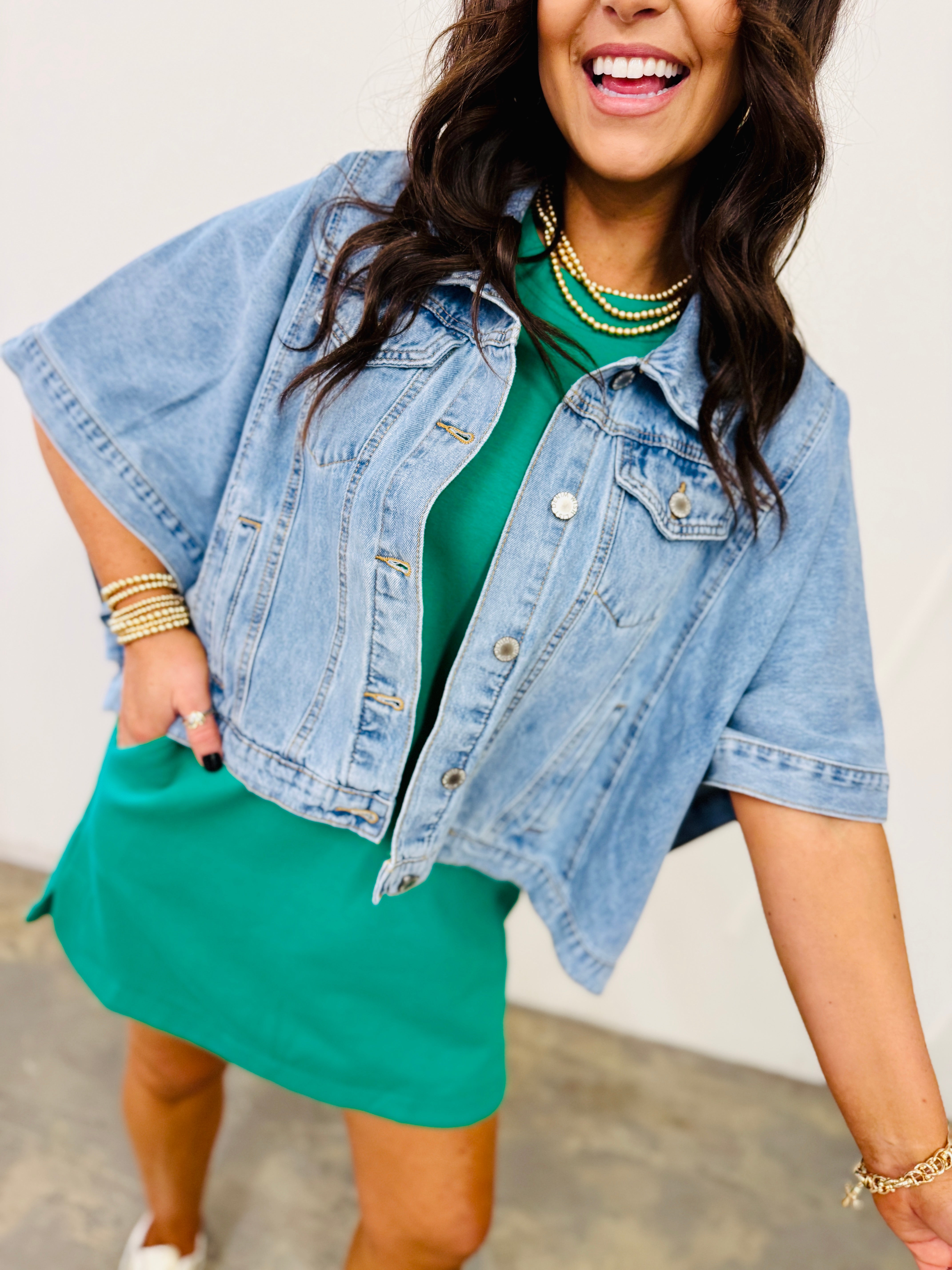 Woman wearing light blue denim Alex jacket with short sleeves over green dress, styled with pearl jewelry.