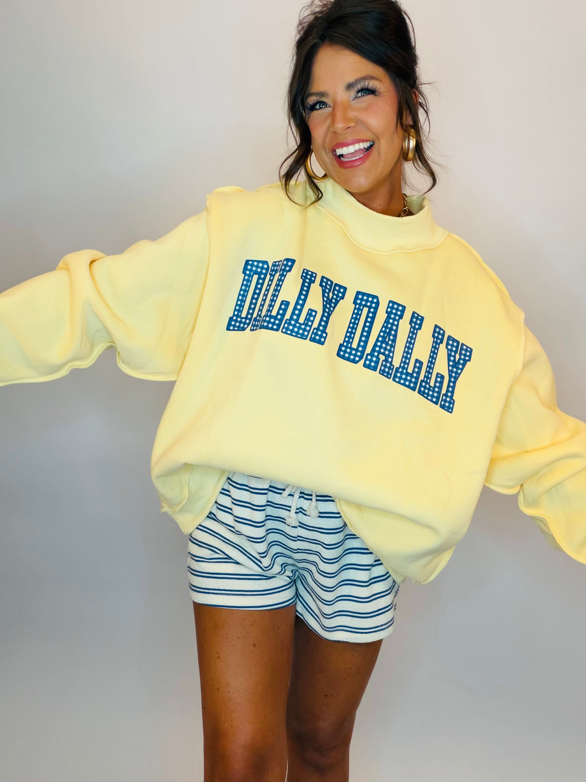 Woman wearing yellow Dilly Dally mock neck sweatshirt with blue polka dot text and blue striped shorts.