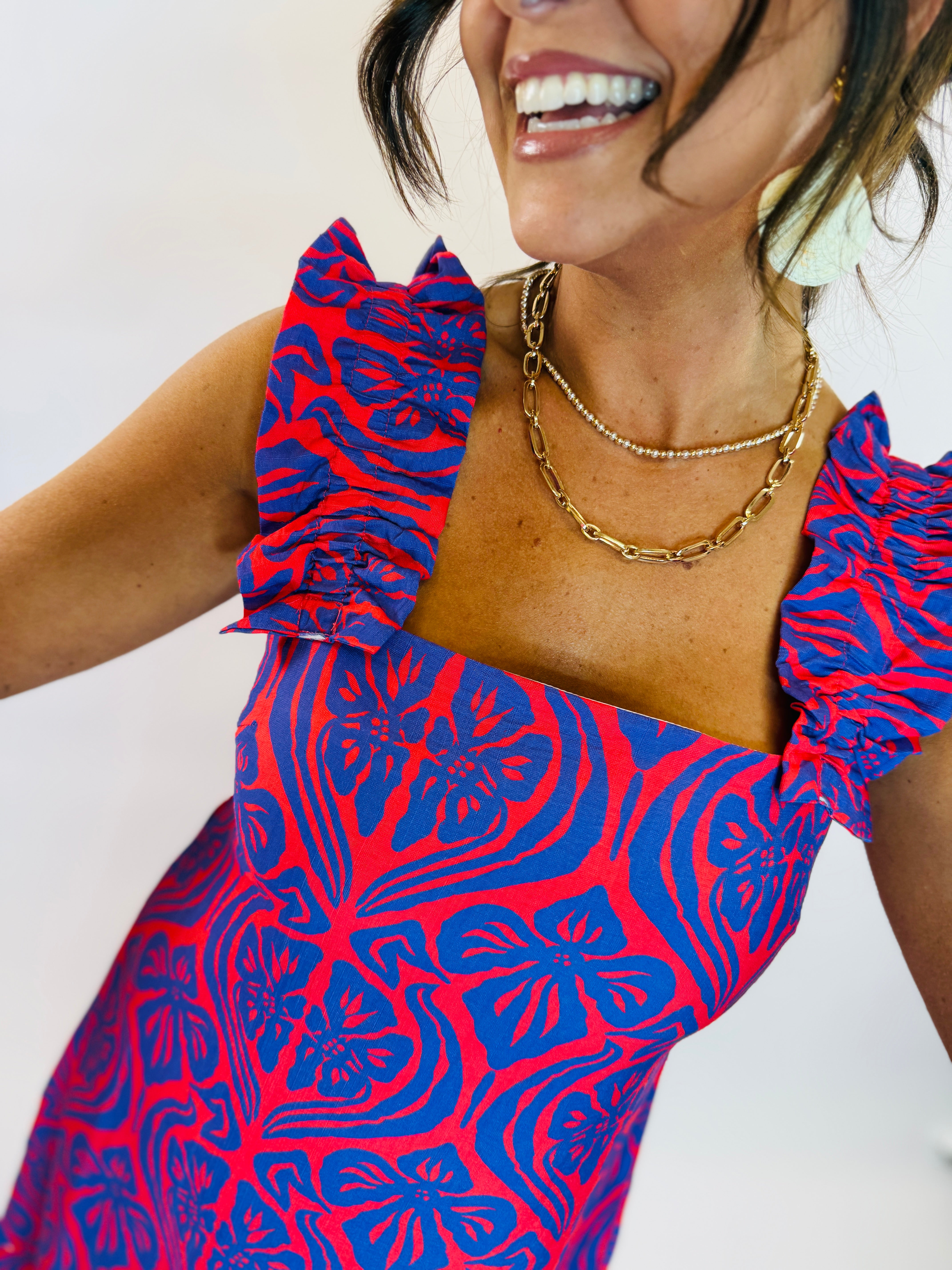 Woman wearing red and blue patterned Ciara dress with ruffle straps and layered gold necklaces, smiling.