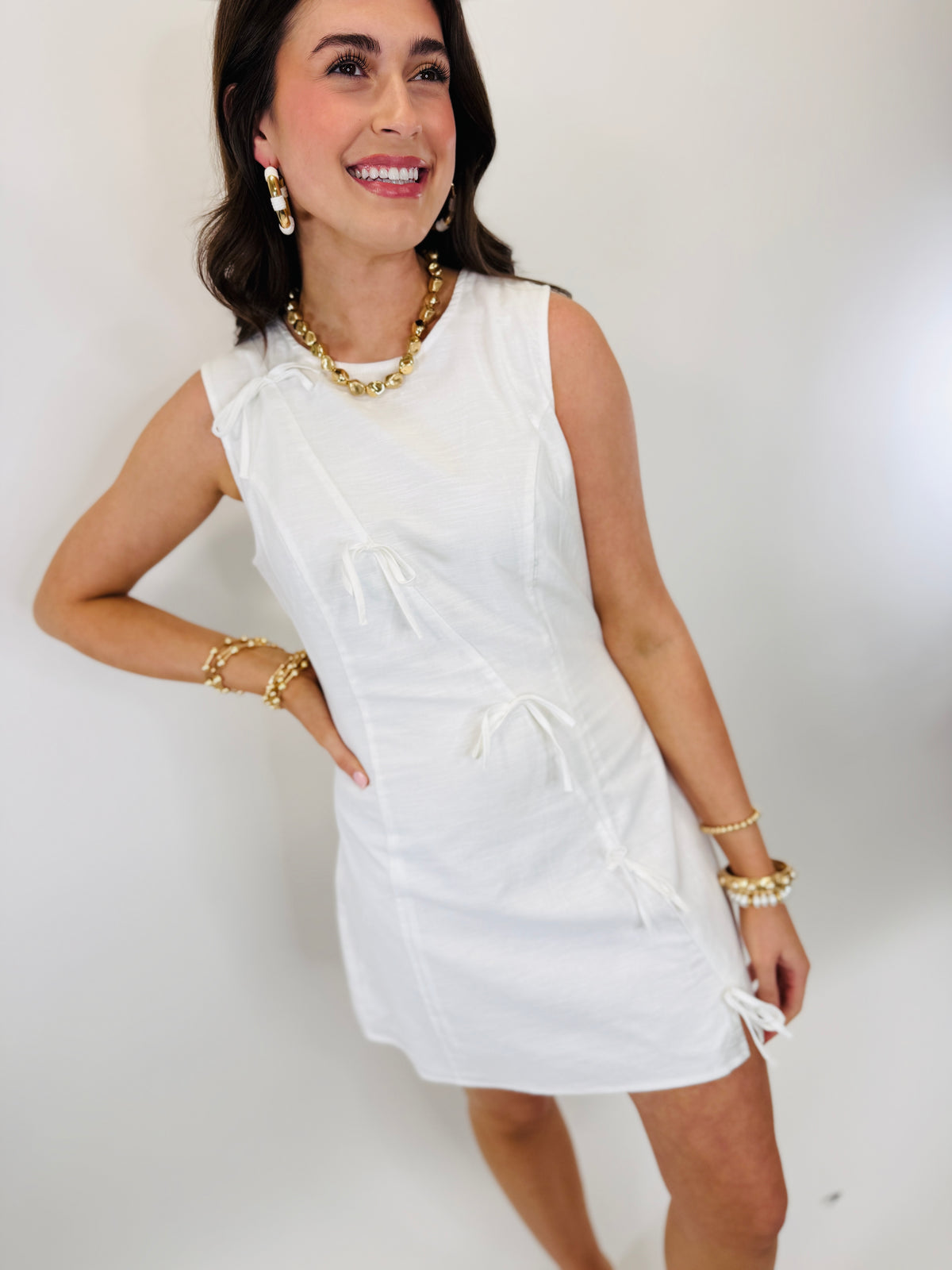 Woman wearing white sleeveless Amanda dress with gold jewelry, smiling against white background