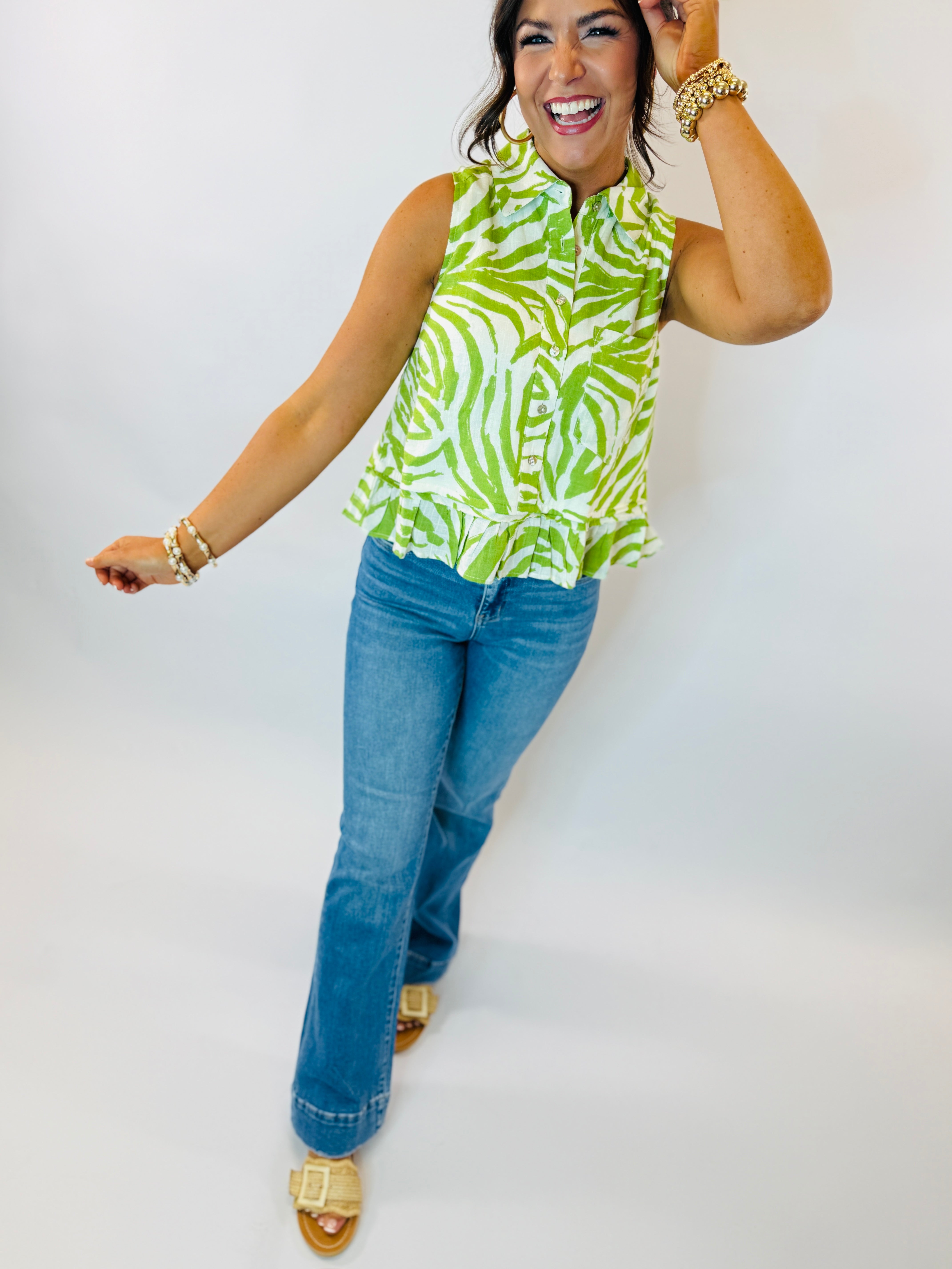 Woman wearing green and white zebra print sleeveless button-up top with blue jeans and gold accessories.