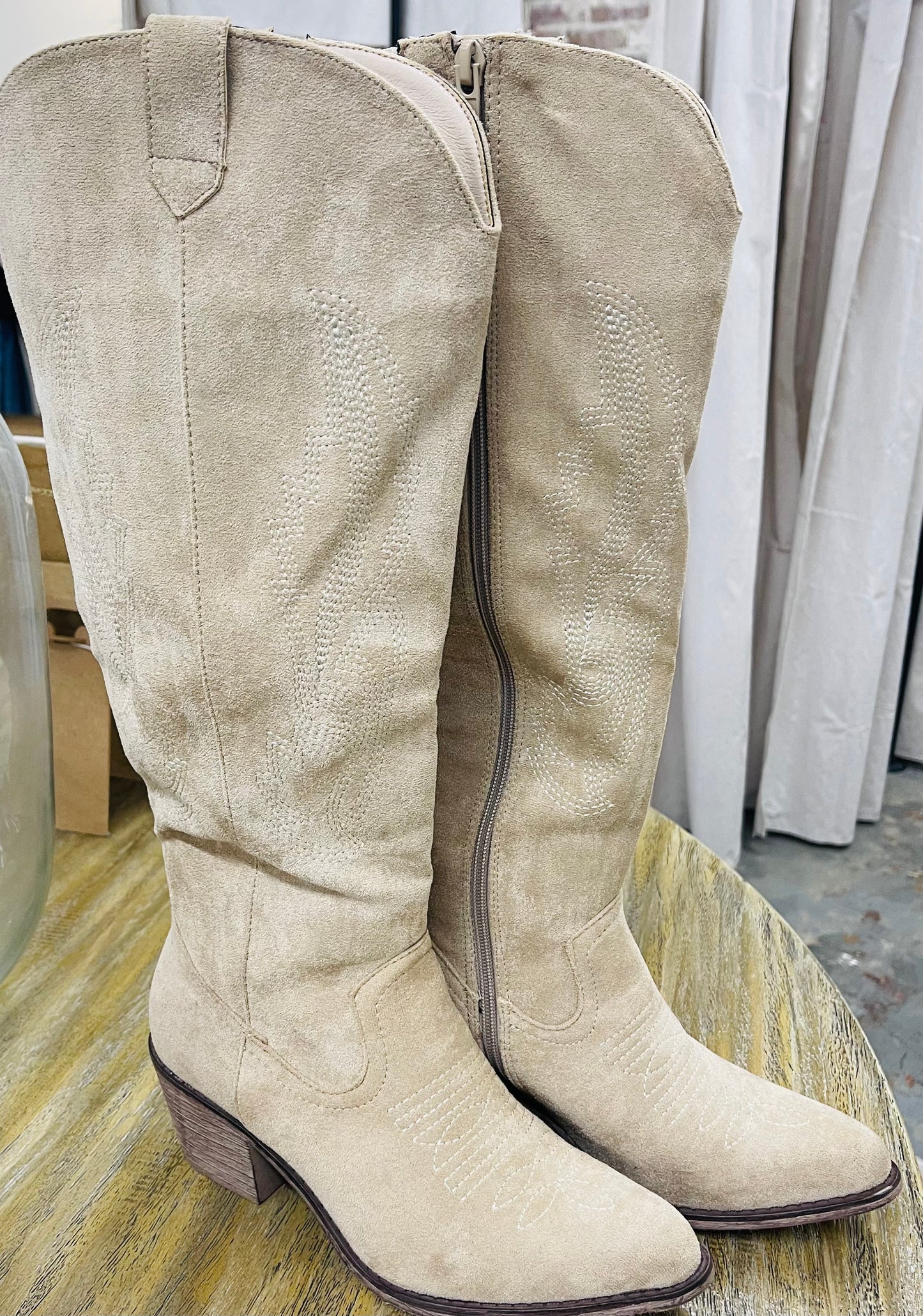 Beige suede WILDER tall boots with decorative stitching, side zipper, and stacked heel on wooden surface.