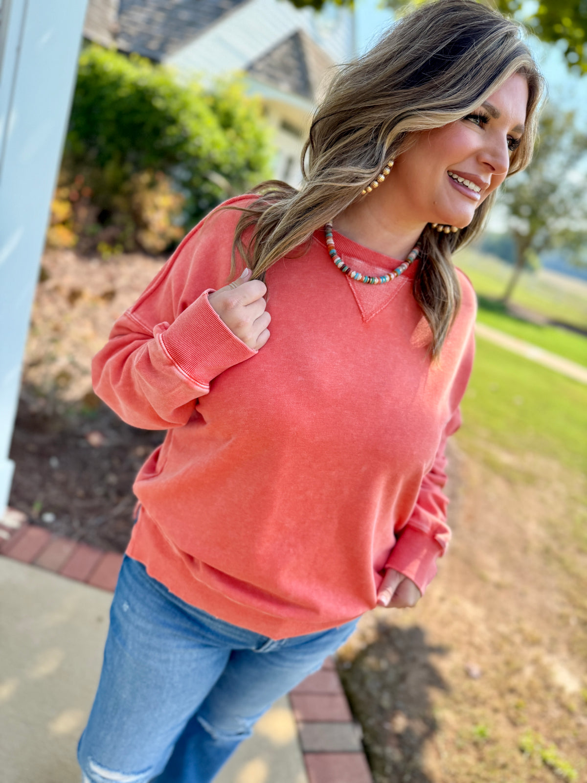 Woman wearing coral pink Danna sweatshirt with blue jeans and colorful beaded necklace, smiling outdoors.
