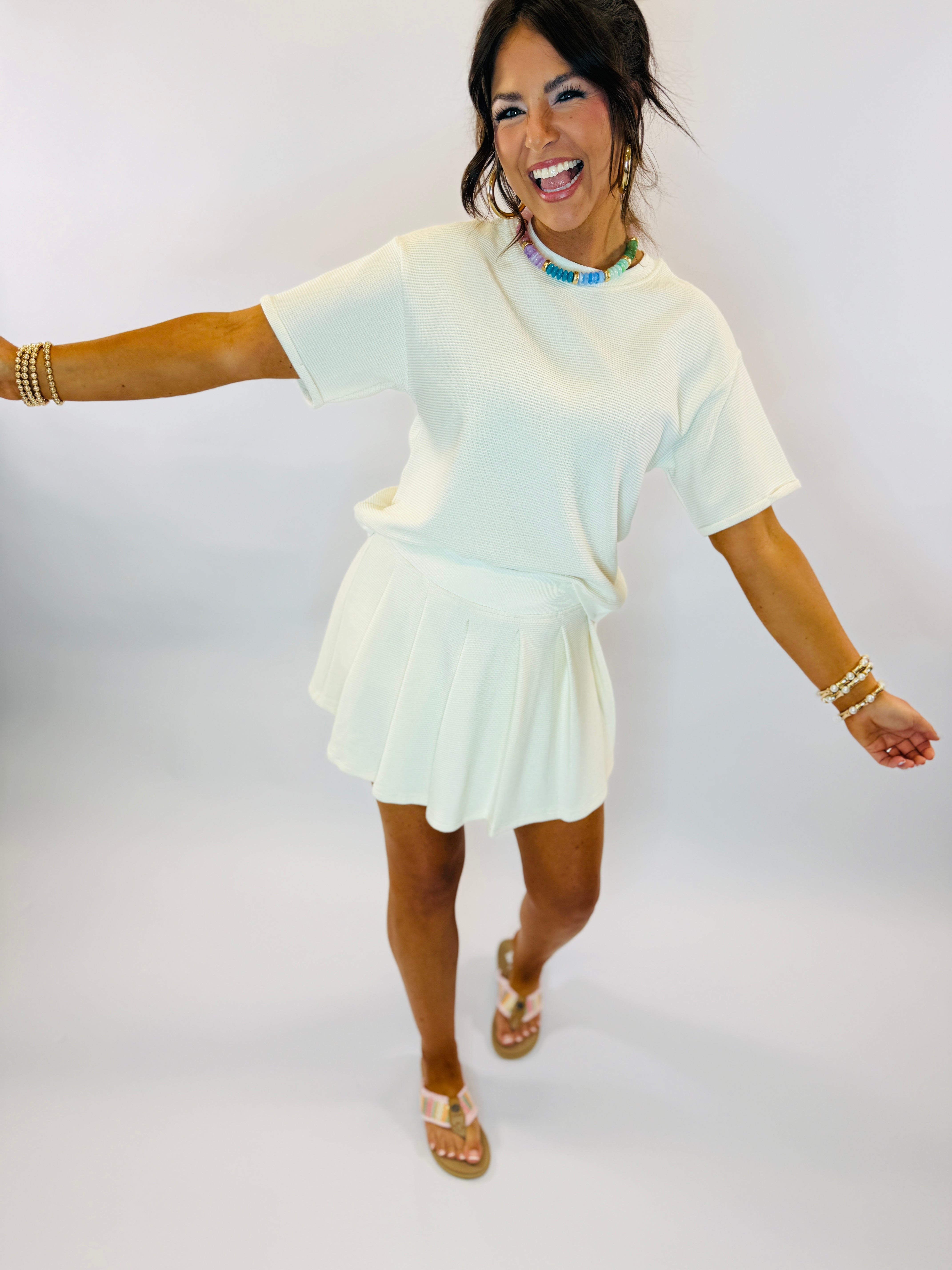 Woman wearing white ribbed short-sleeve top and matching pleated mini skort, accessorized with colorful beaded necklace an...