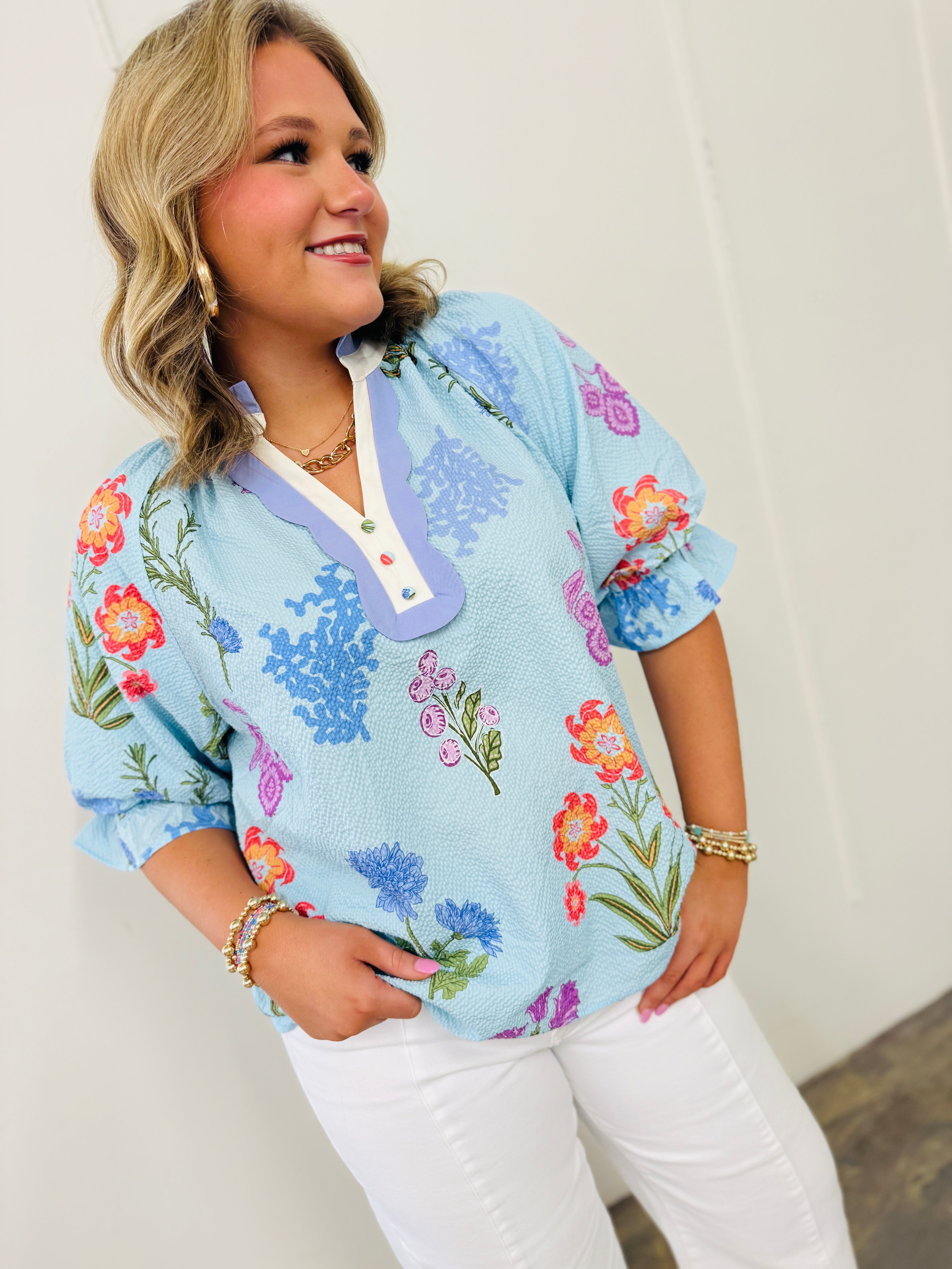 Woman wearing light blue floral blouse with colorful flower pattern and purple collar trim, paired with white pants.