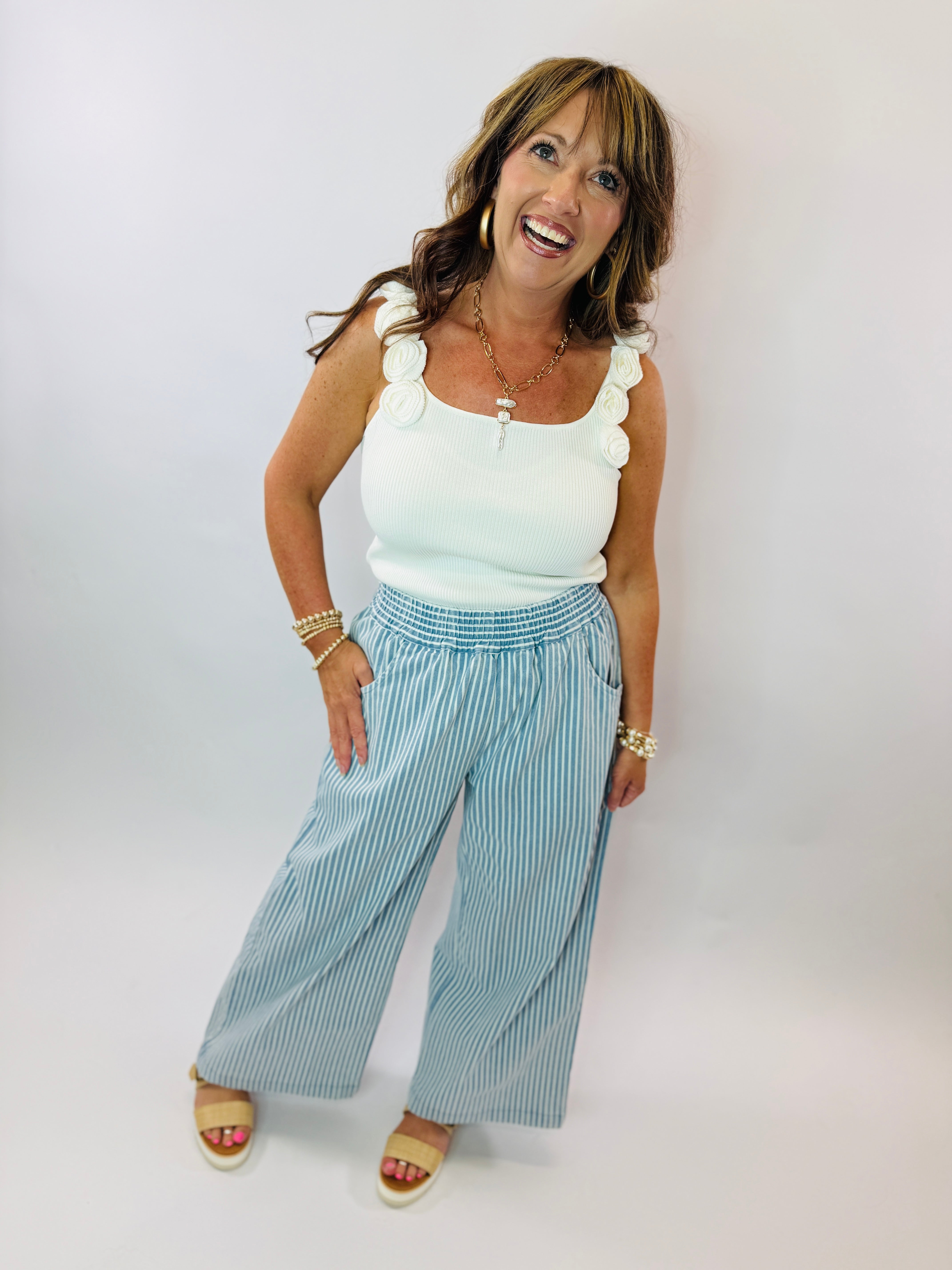 Woman wearing white ribbed sleeveless sweater tank with scalloped straps, paired with blue striped wide-leg pants and sand...