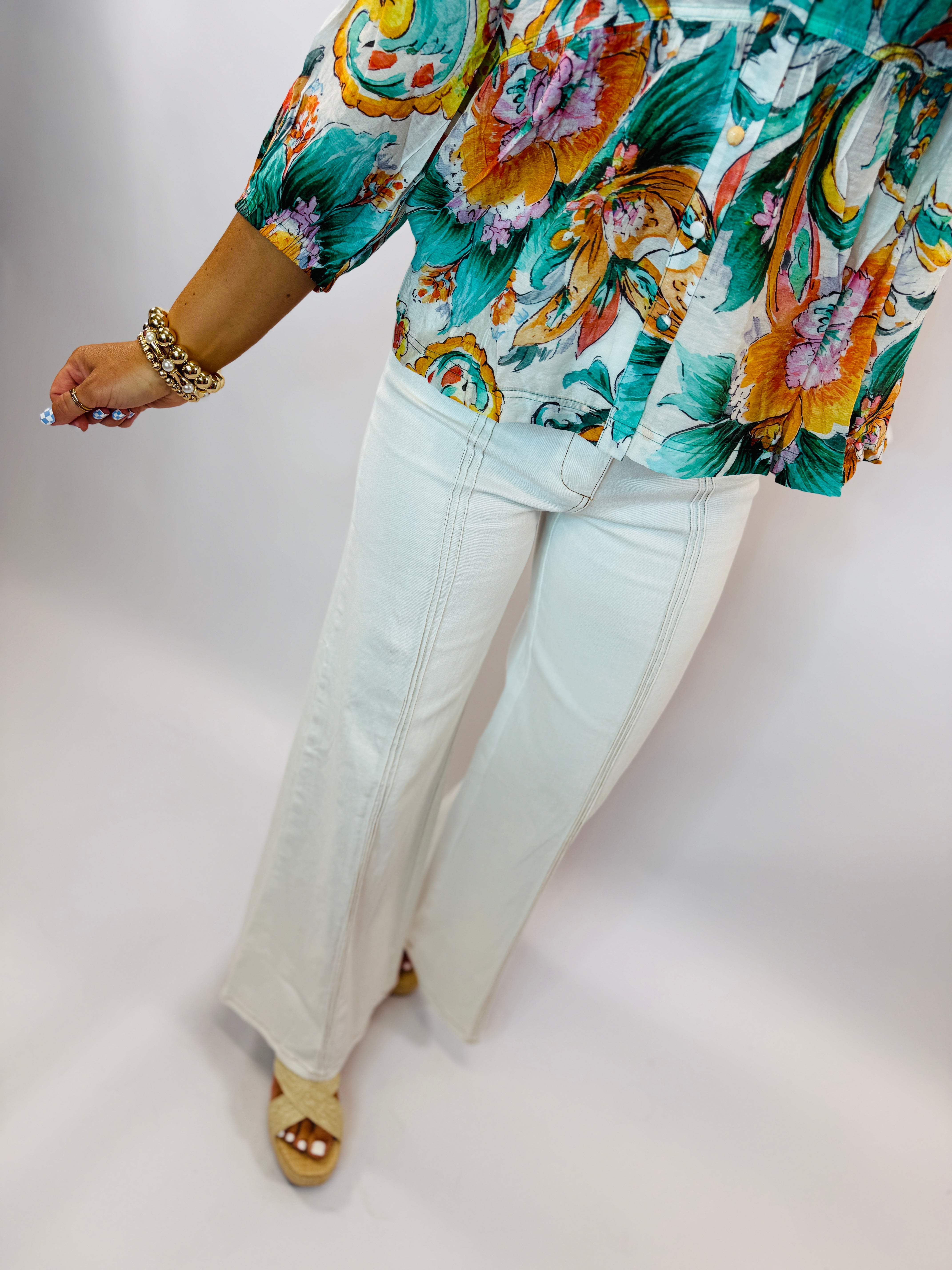 Woman wearing colorful tropical floral print top with white flare jeans and gold chain bracelet
