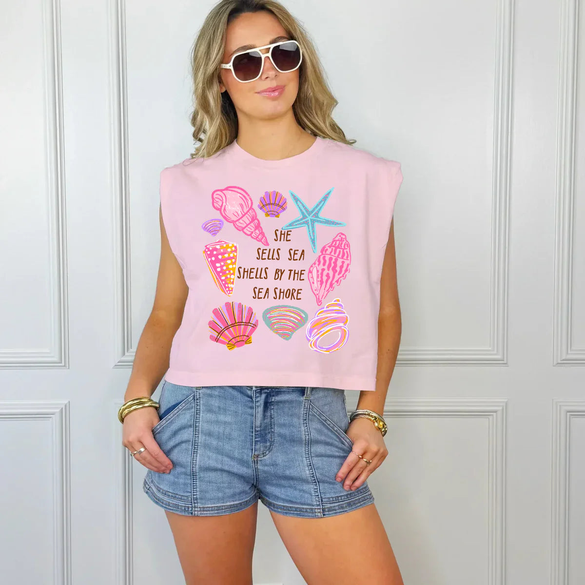 Woman wearing pink sleeveless tank top with colorful seashell graphics and text, paired with denim shorts.