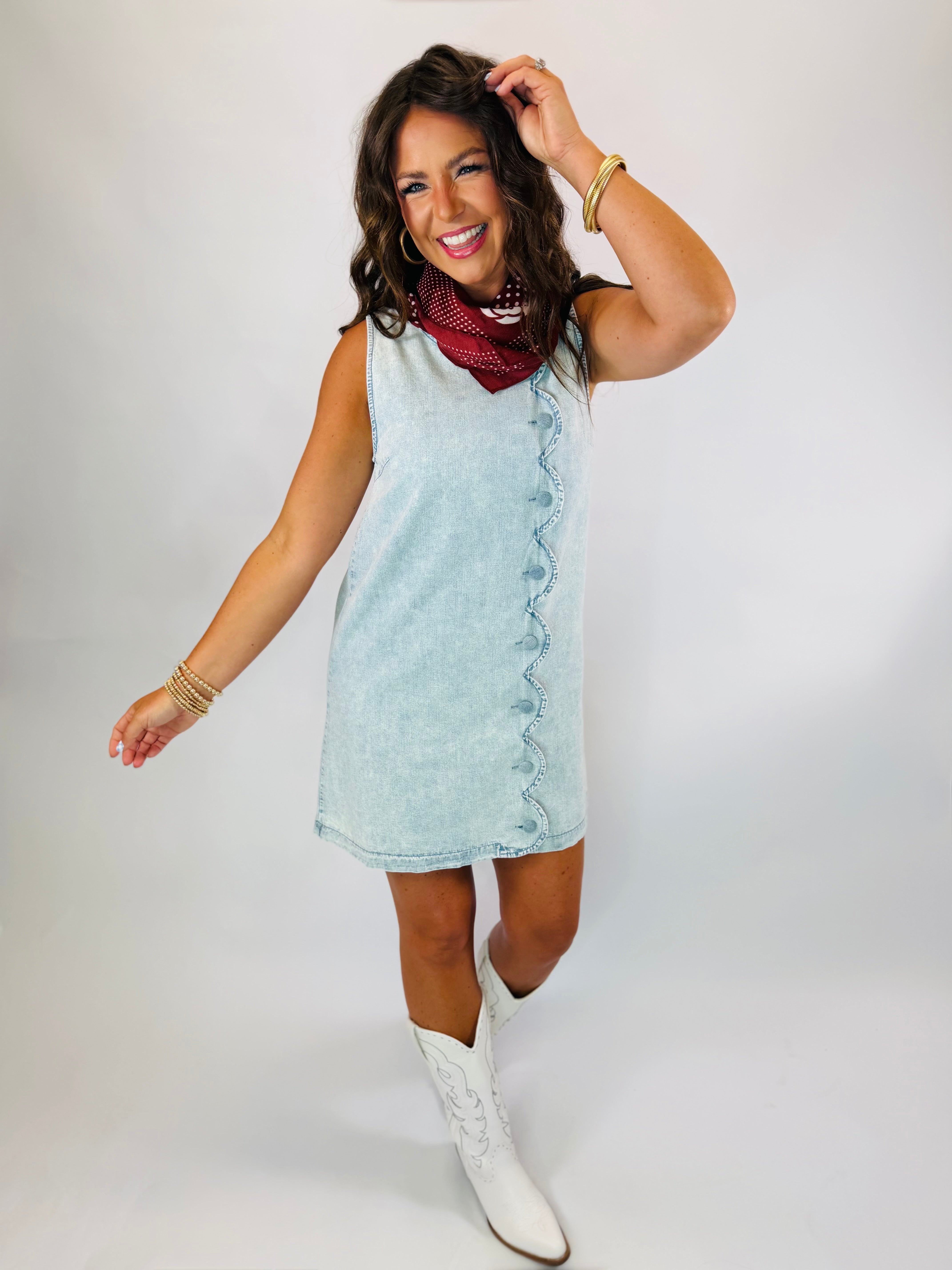 Woman wearing light wash sleeveless denim mini dress with scalloped button detail, styled with white cowboy boots and red ...