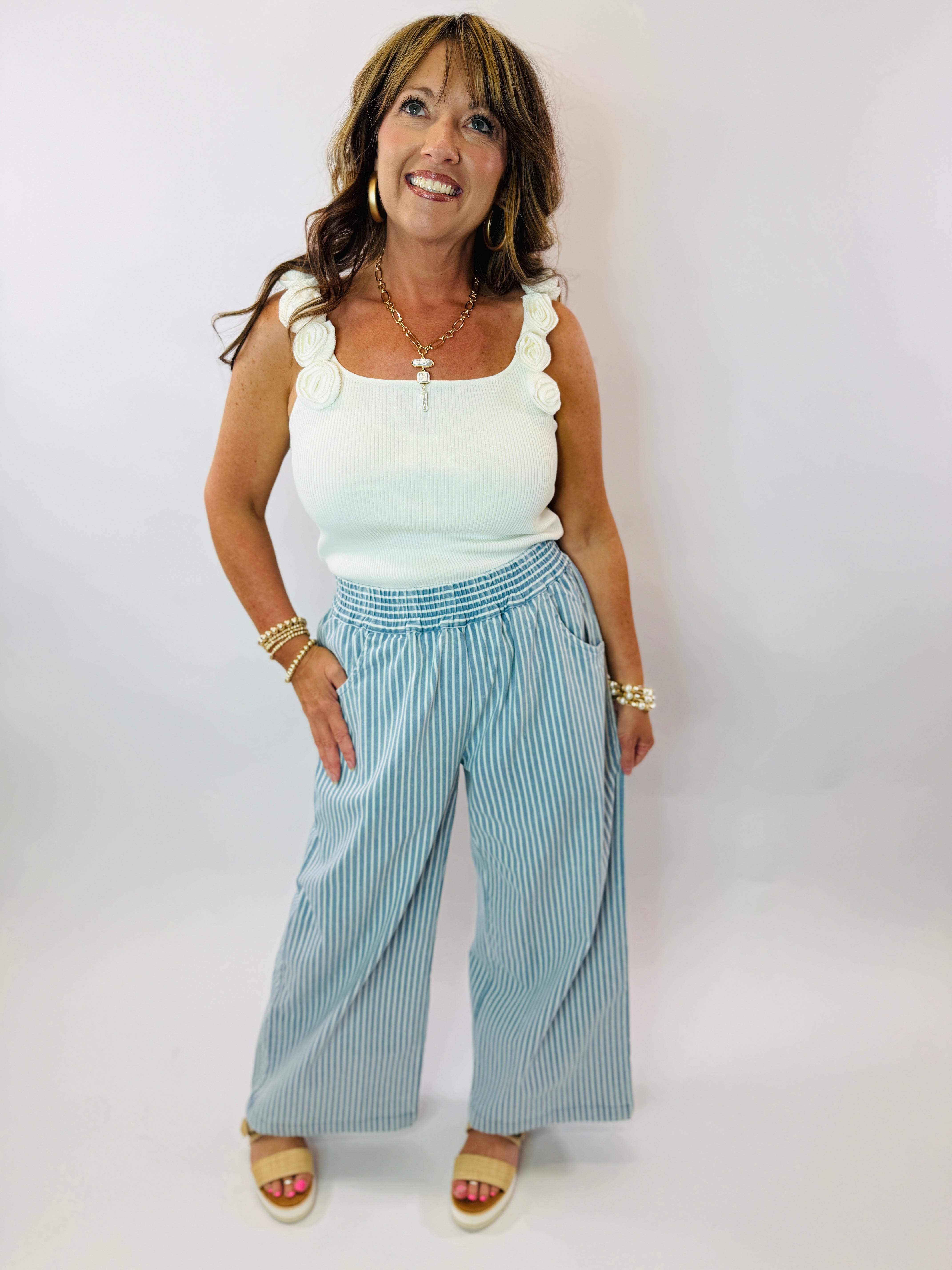Woman wearing white ribbed tank top with ruffled straps, blue striped wide-leg pants, and gold jewelry