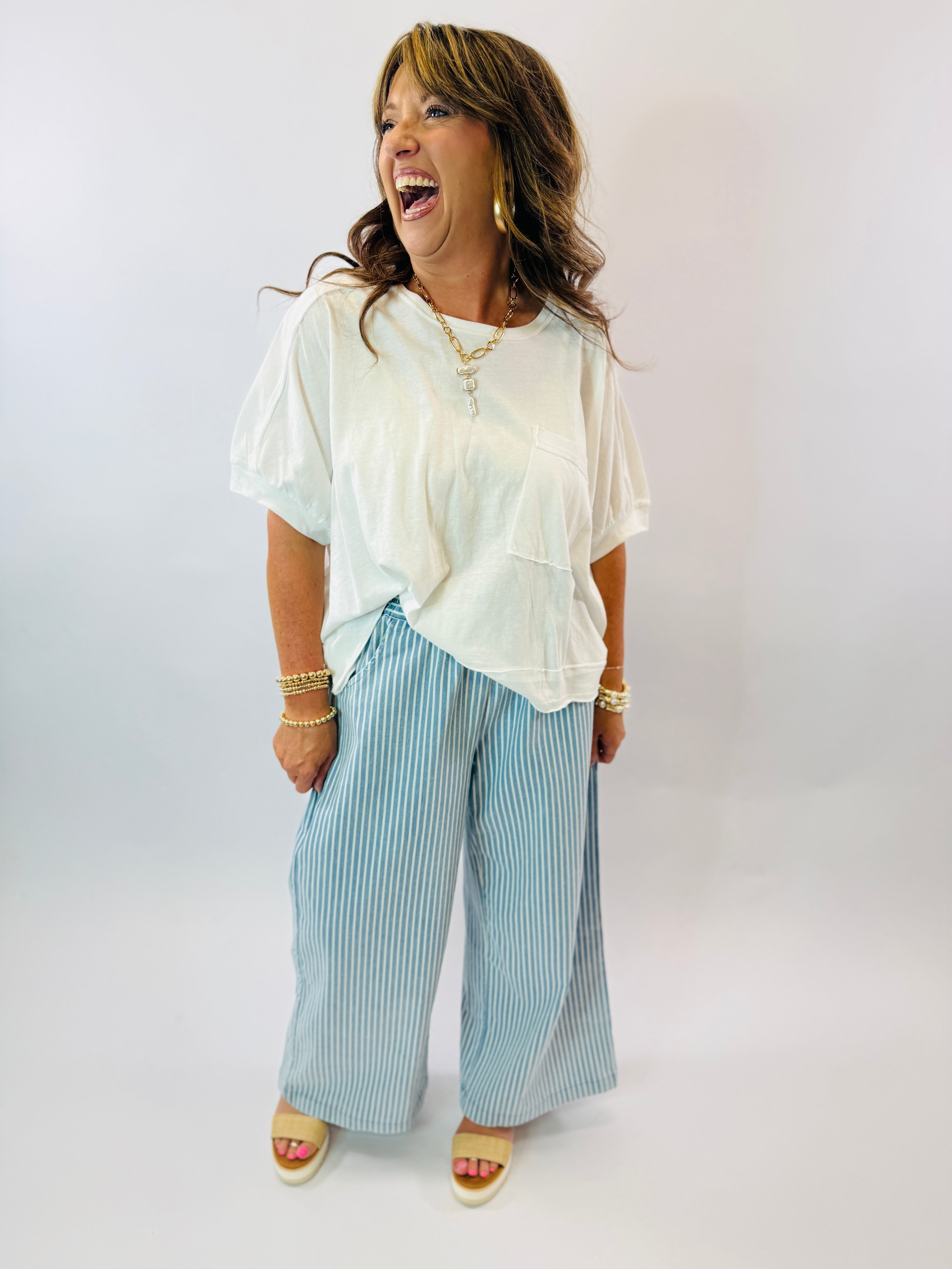 Woman wearing white oversized t-shirt and blue striped wide leg pants, laughing and accessorized with gold jewelry.