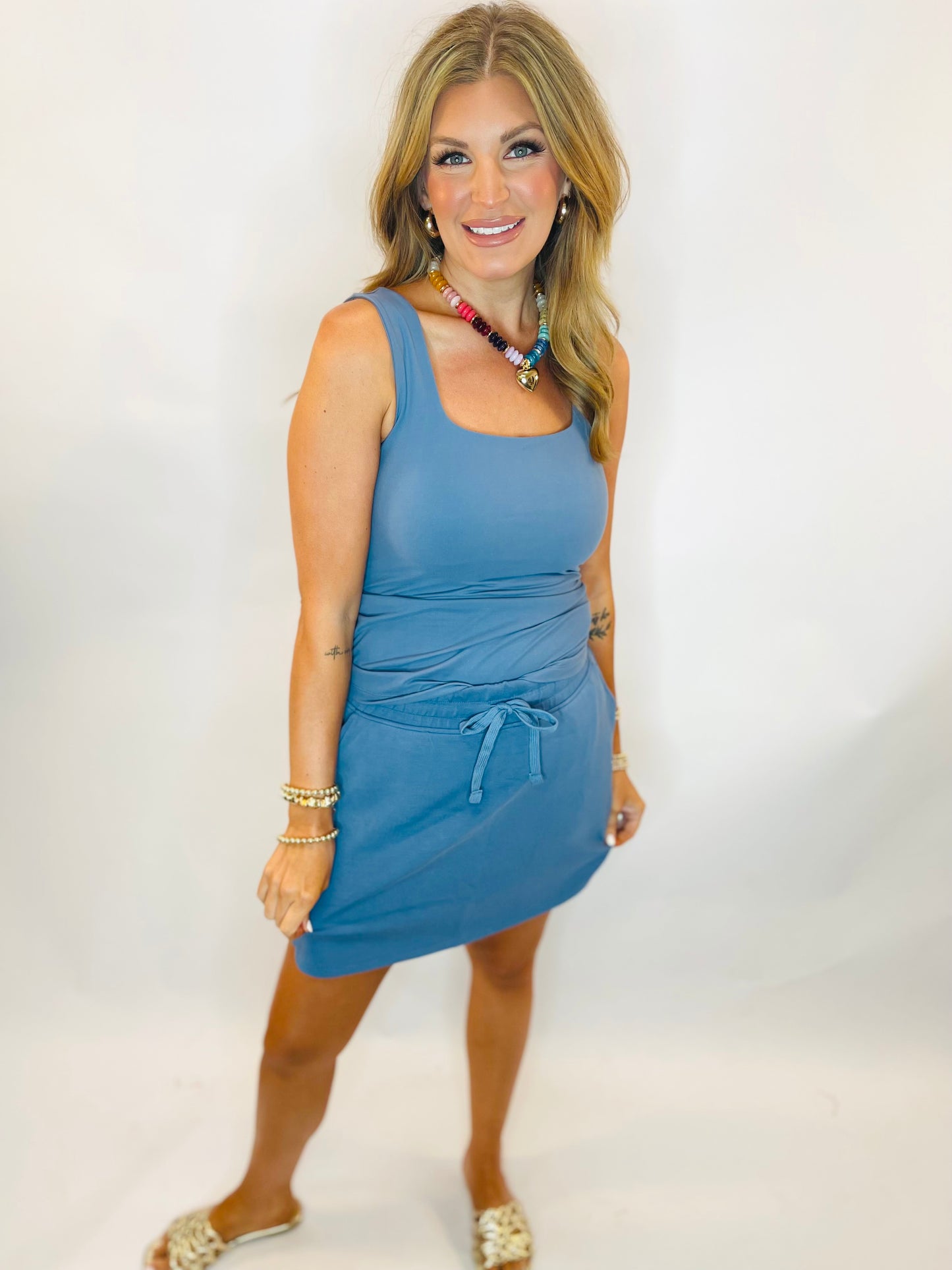 Woman wearing a sleeveless blue ruched mini dress with drawstring waist detail and square neckline.
