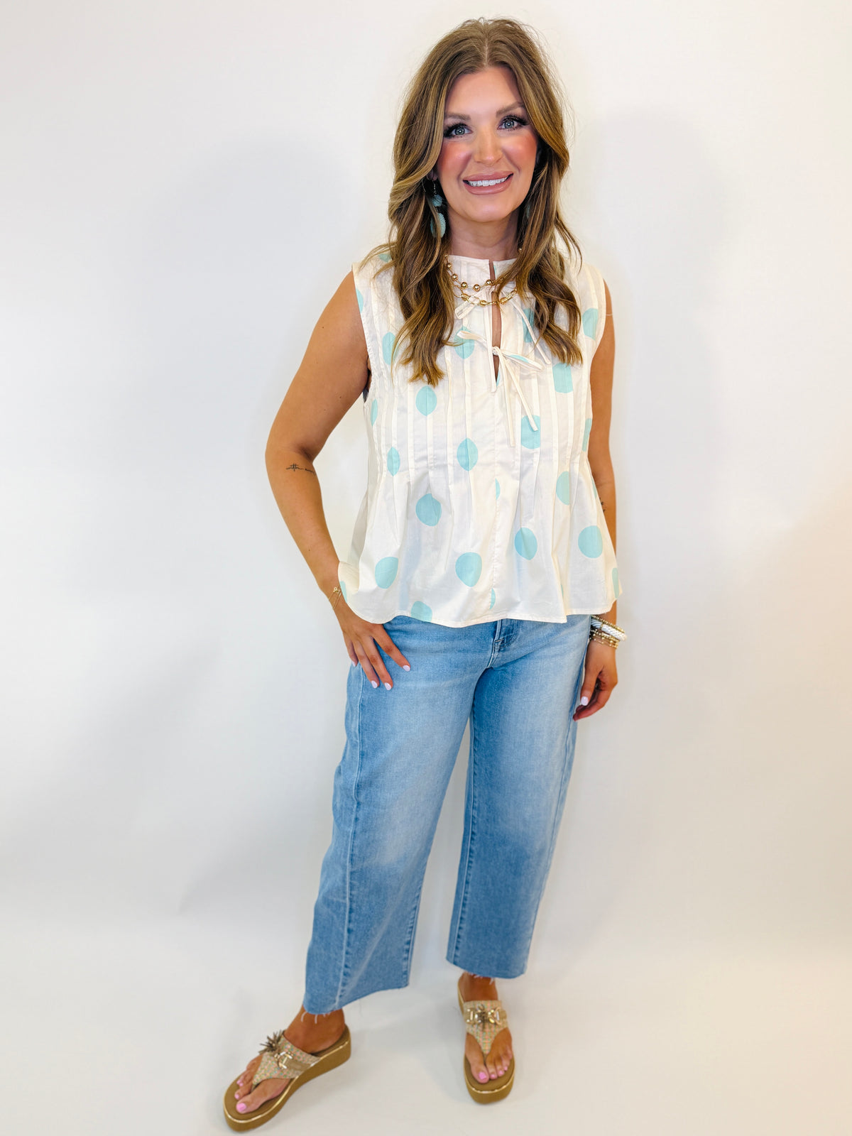 Woman wearing white sleeveless top with blue polka dots, cropped jeans, and gold sandals against white background.