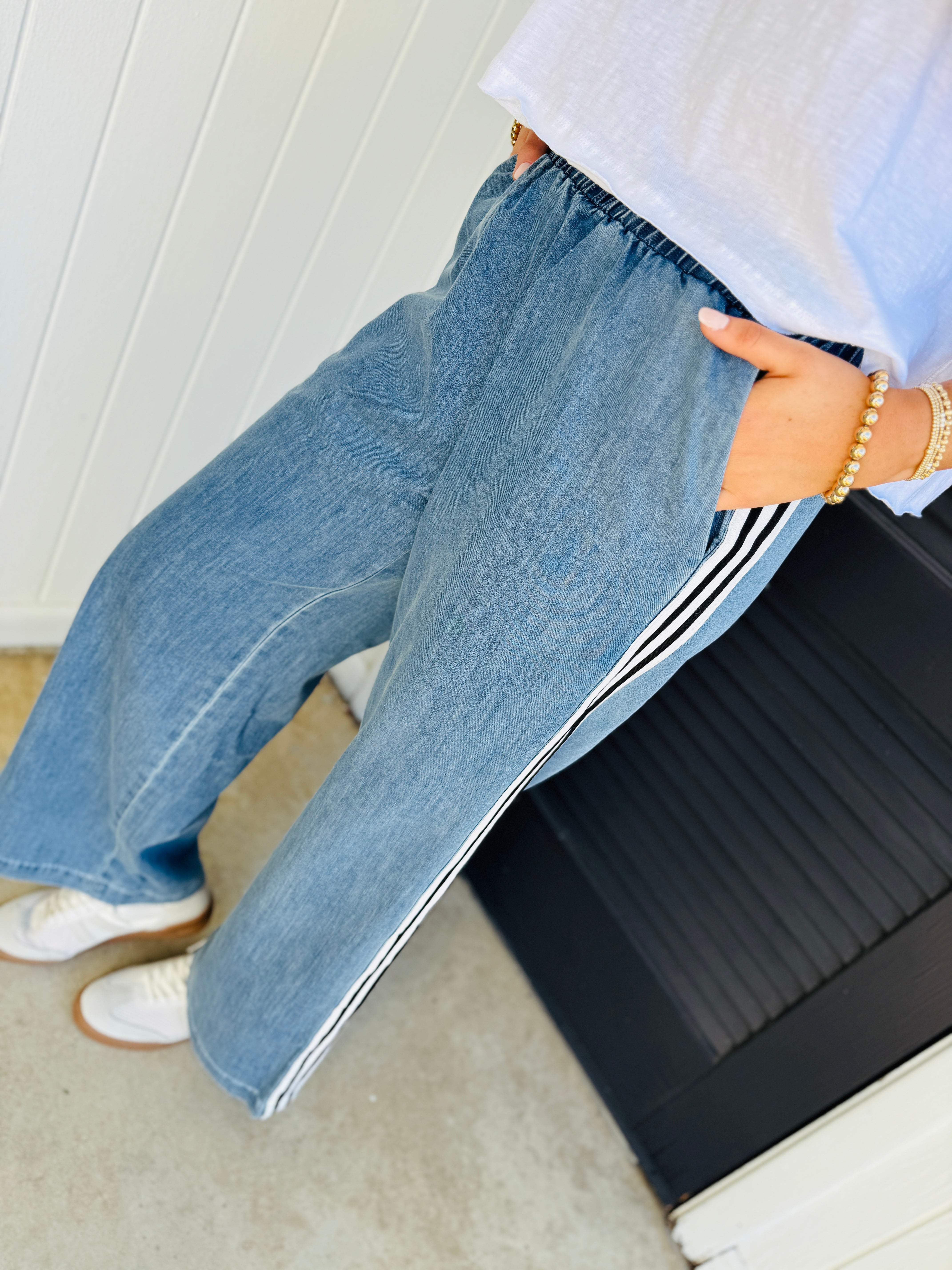 Light blue denim relaxed pants with white side stripes, worn with white sneakers and pearl bracelet.