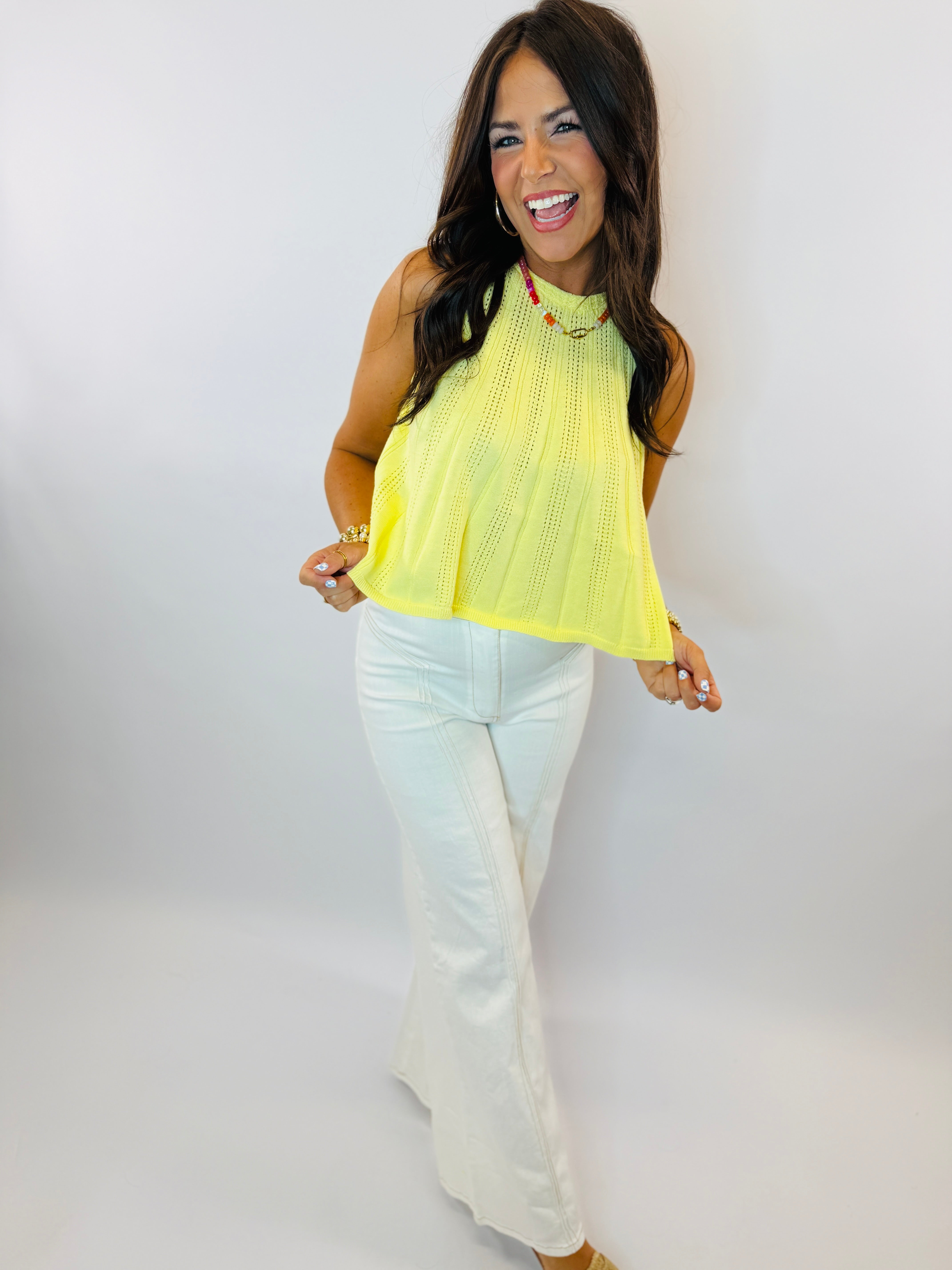Woman wearing bright yellow sleeveless knit top with white pants, smiling against light background