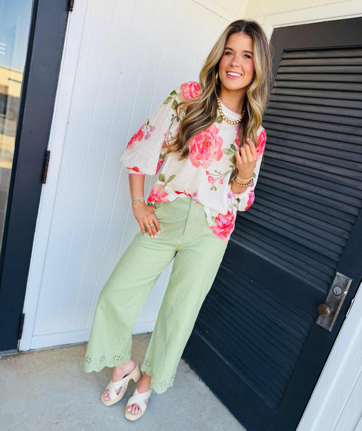 Woman wearing white floral top with pink roses and green pants, styled with pearl jewelry and wedge sandals.