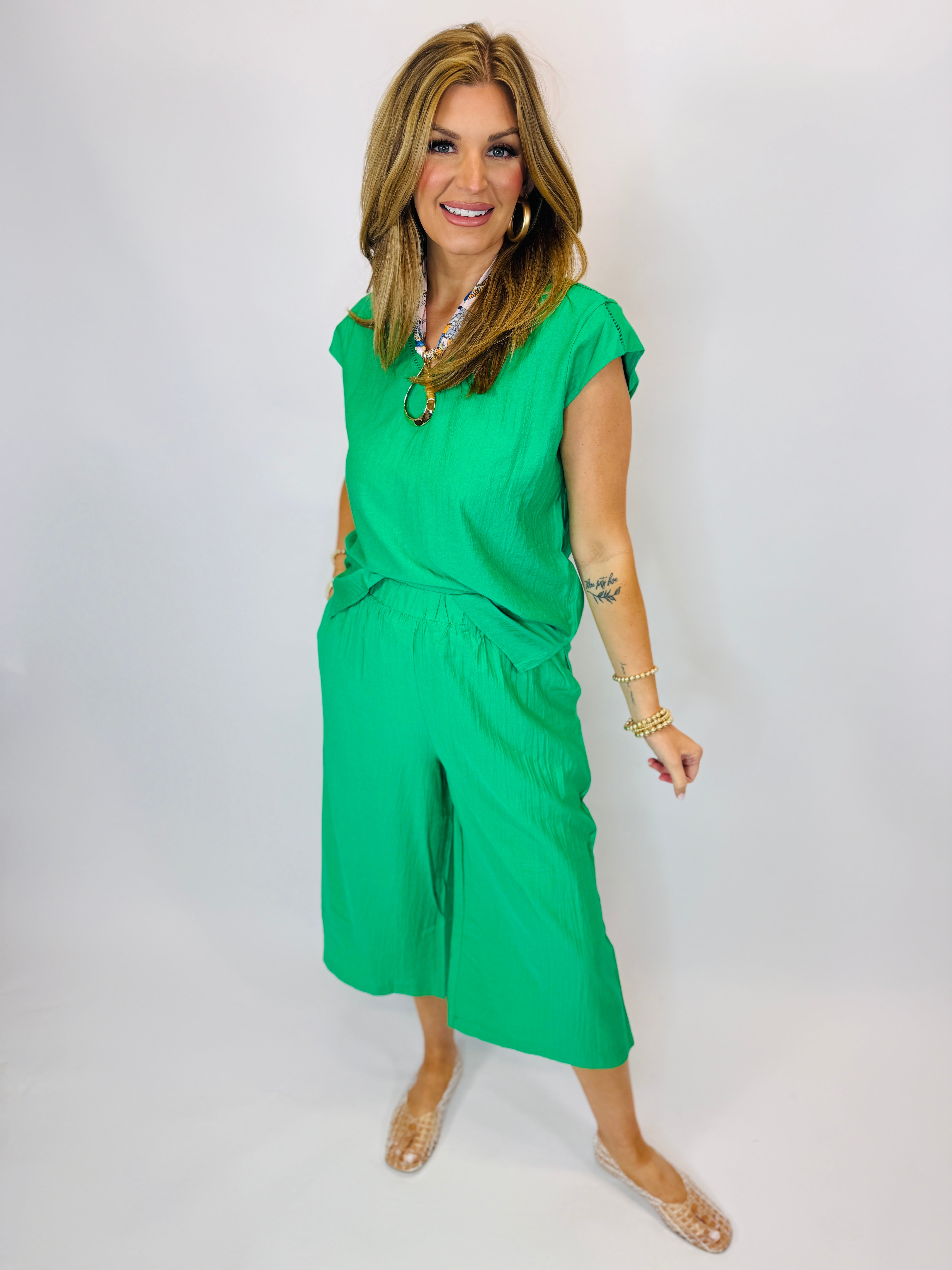 Woman wearing bright green short-sleeve top and matching wide-leg cropped pants with gold jewelry and sandals.