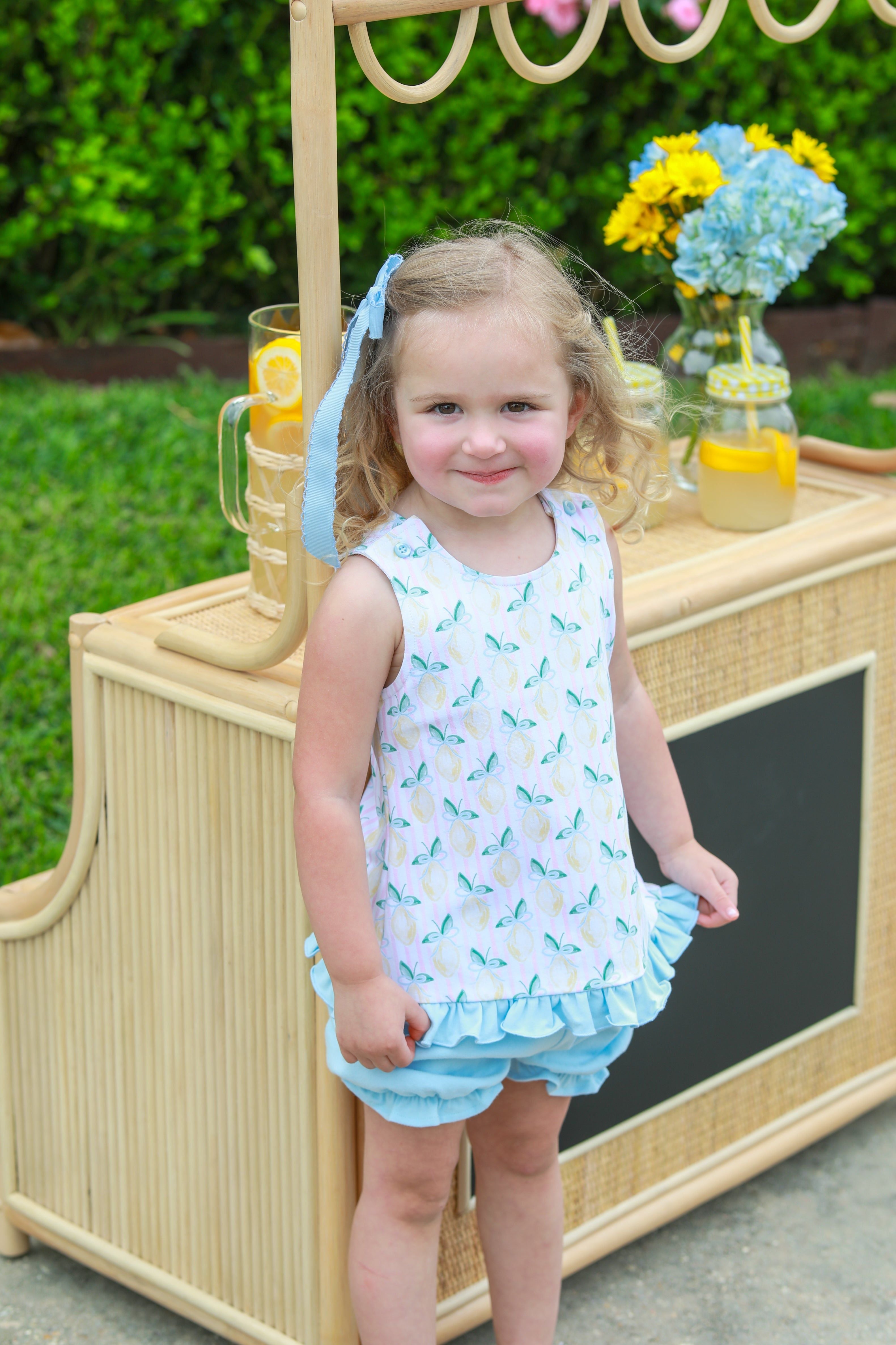 Young girl wearing white sleeveless top with blue pattern and light blue ruffled bloomers, standing by wooden lemonade stand.