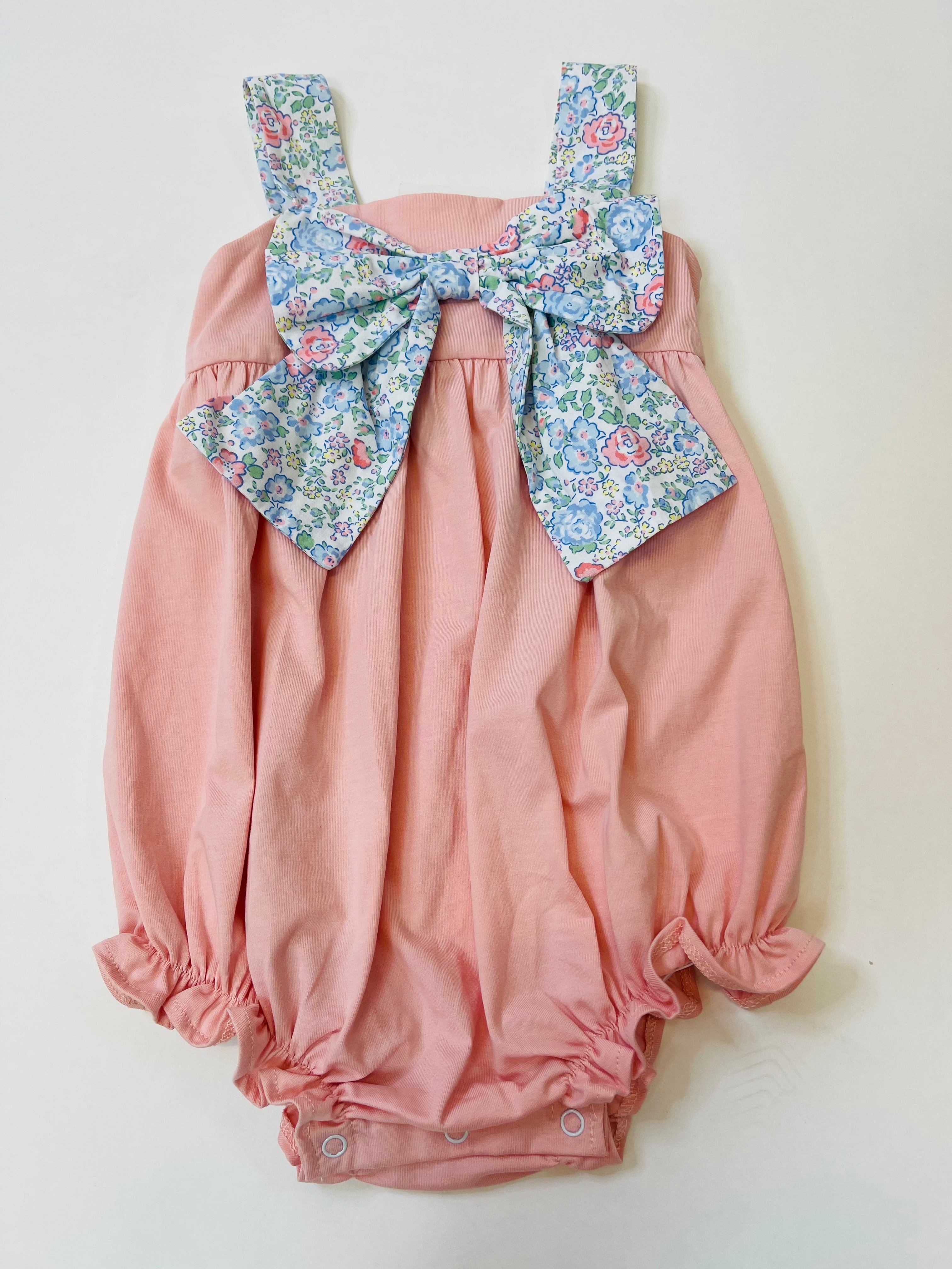 Pink baby bubble romper with floral bow detail and shoulder straps, featuring snap closures at bottom.