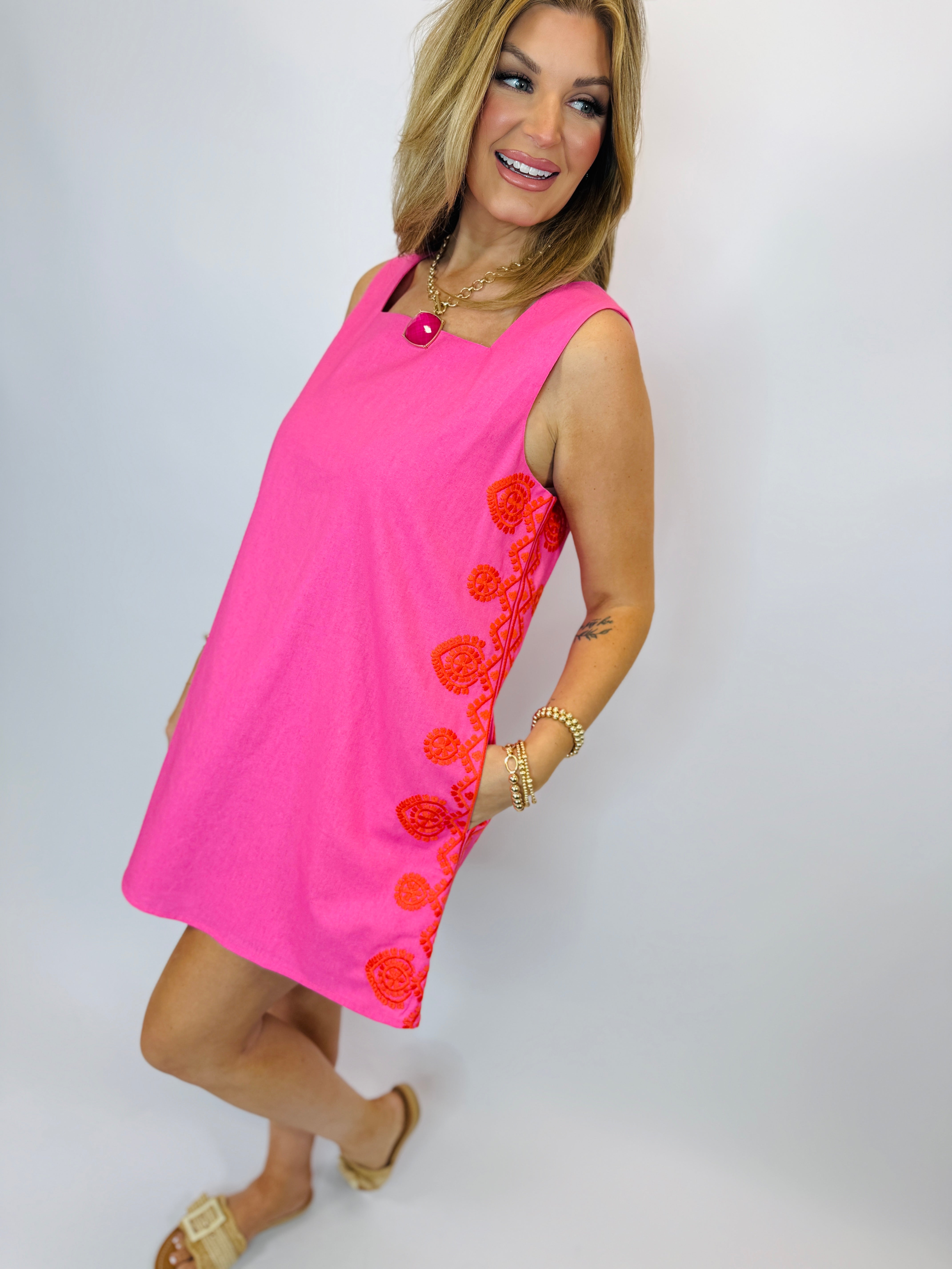 Woman wearing bright pink sleeveless Marlene dress with red floral embroidery along the side, styled with gold jewelry.