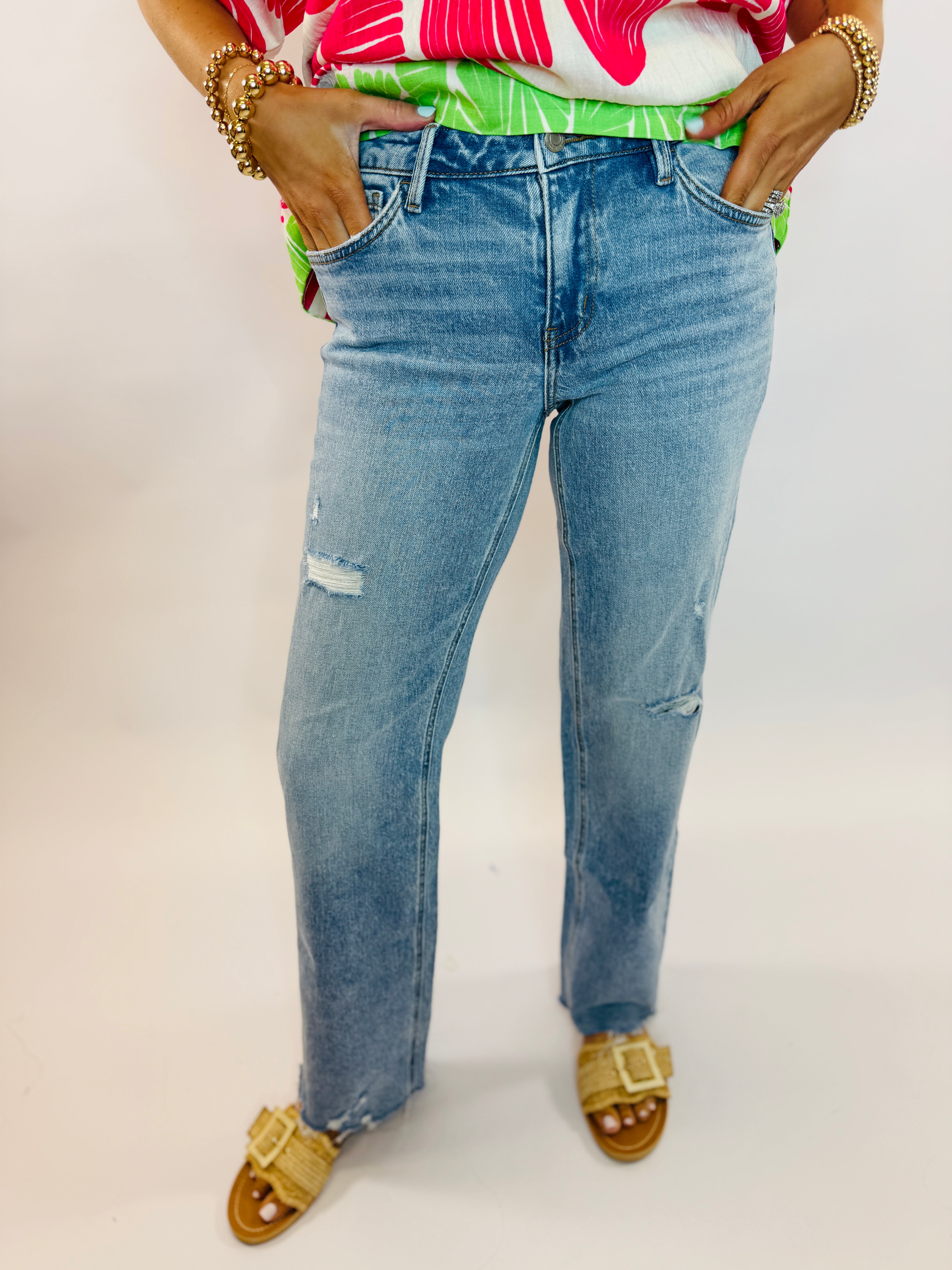 Woman wearing light blue distressed Lena Denim jeans with ripped details, colorful top, and woven sandals.