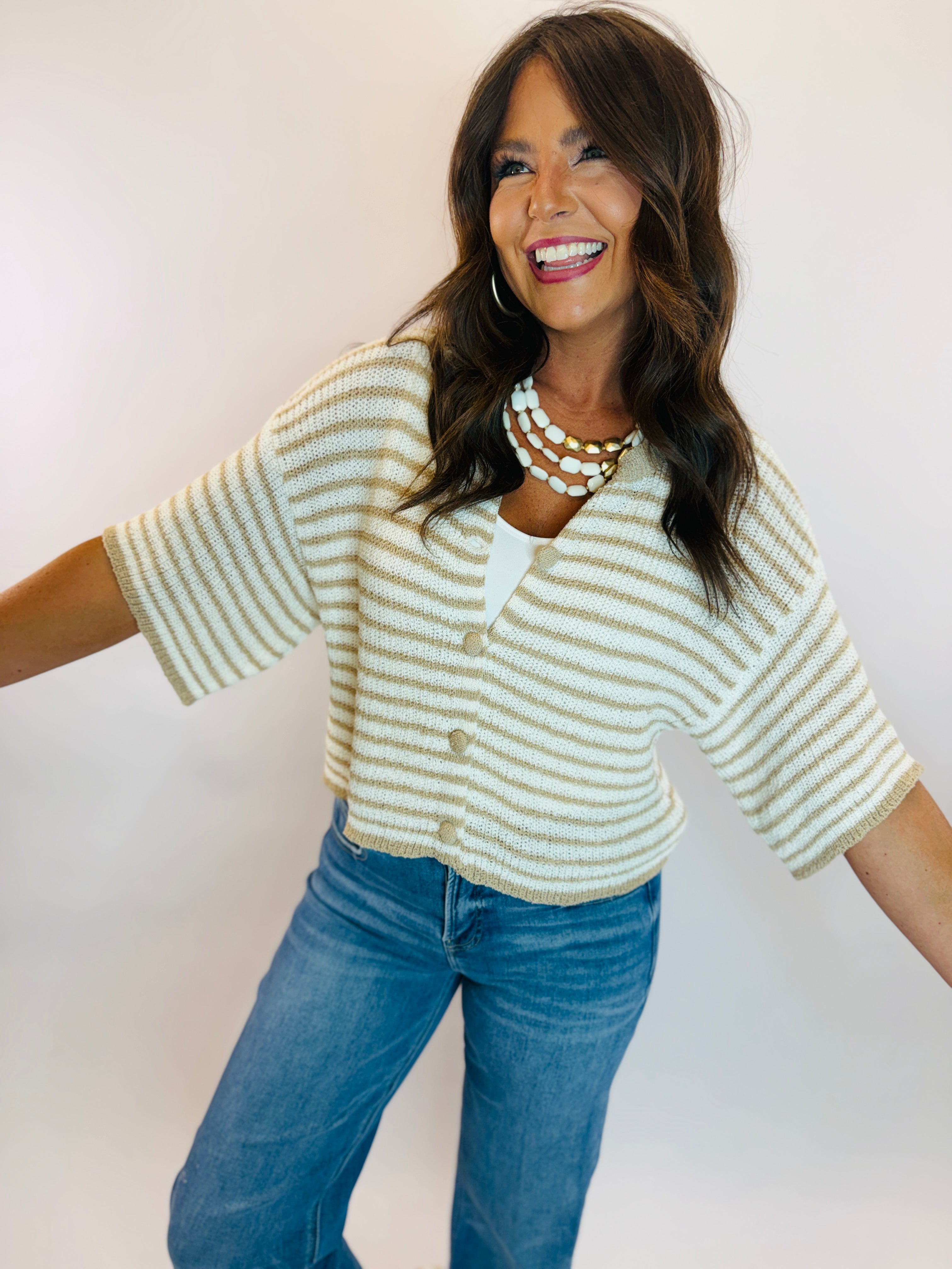 Woman wearing Vivian Top, a cream and tan striped short-sleeve knit cardigan with gold buttons, paired with blue jeans.
