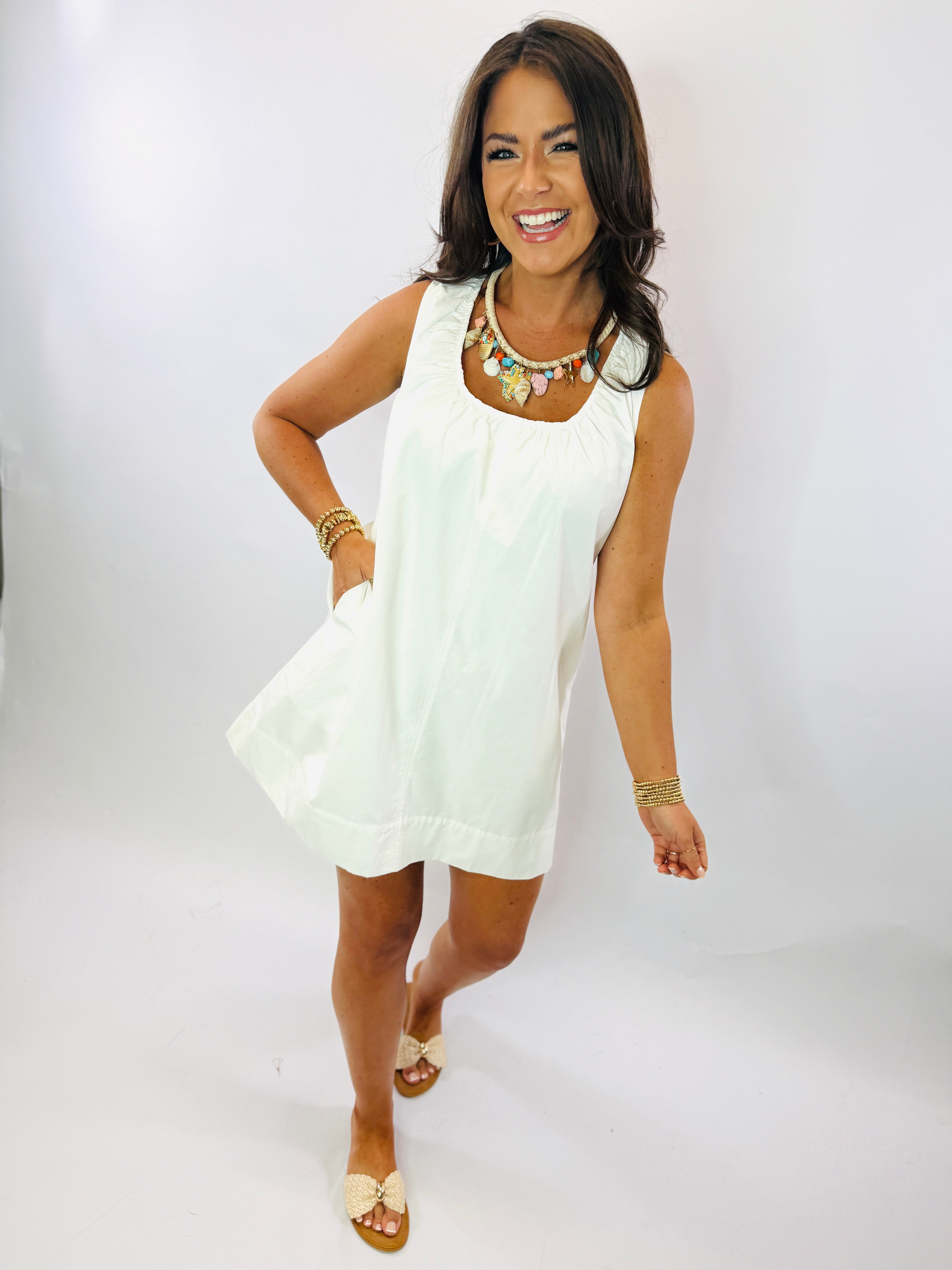 Woman wearing white sleeveless Katherine dress with statement necklace and gold sandals, smiling with hand on hip.