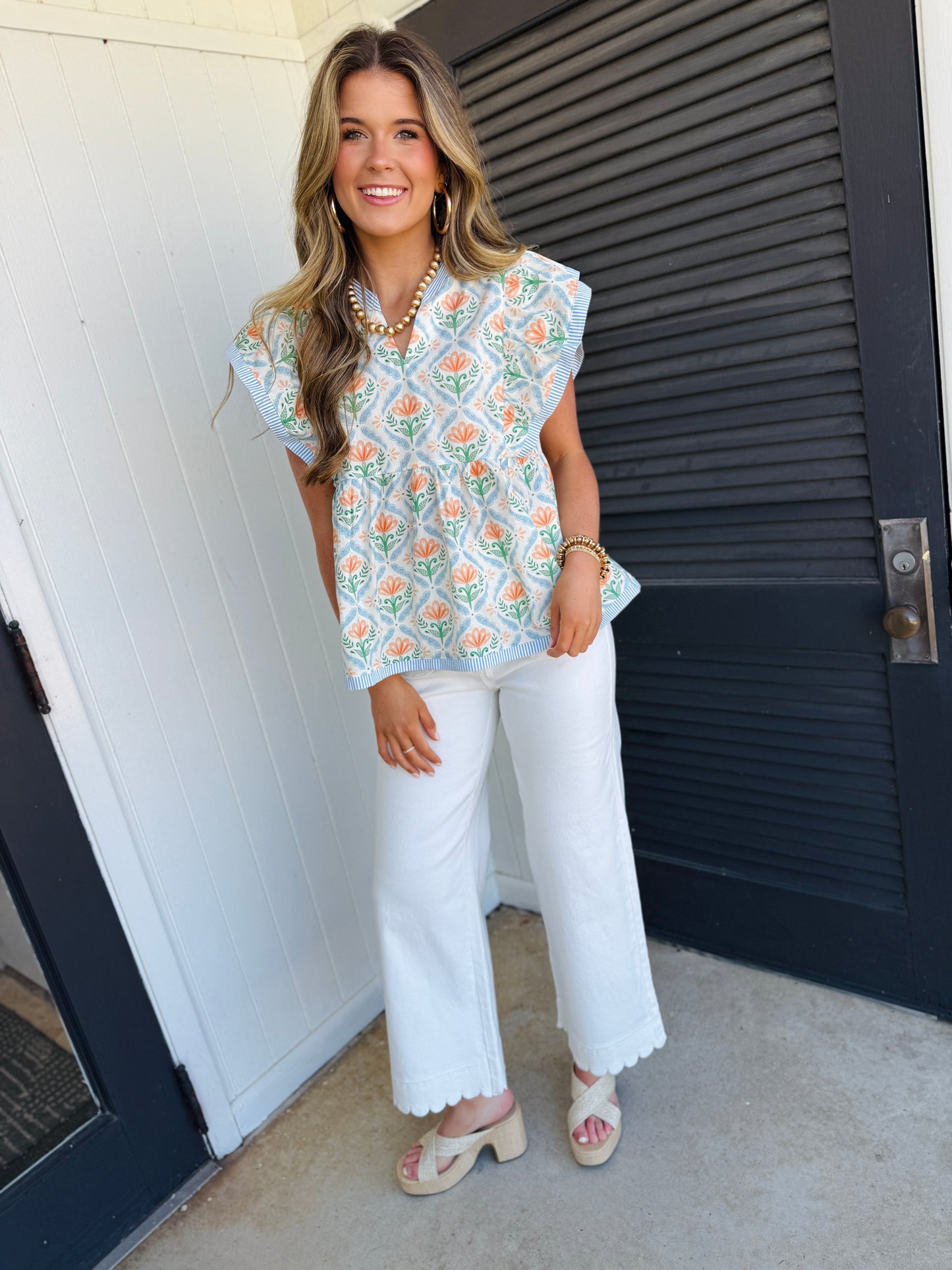 Woman wearing floral patterned Ella Everyday Turner Top with white pants and pearl necklace, smiling outdoors.