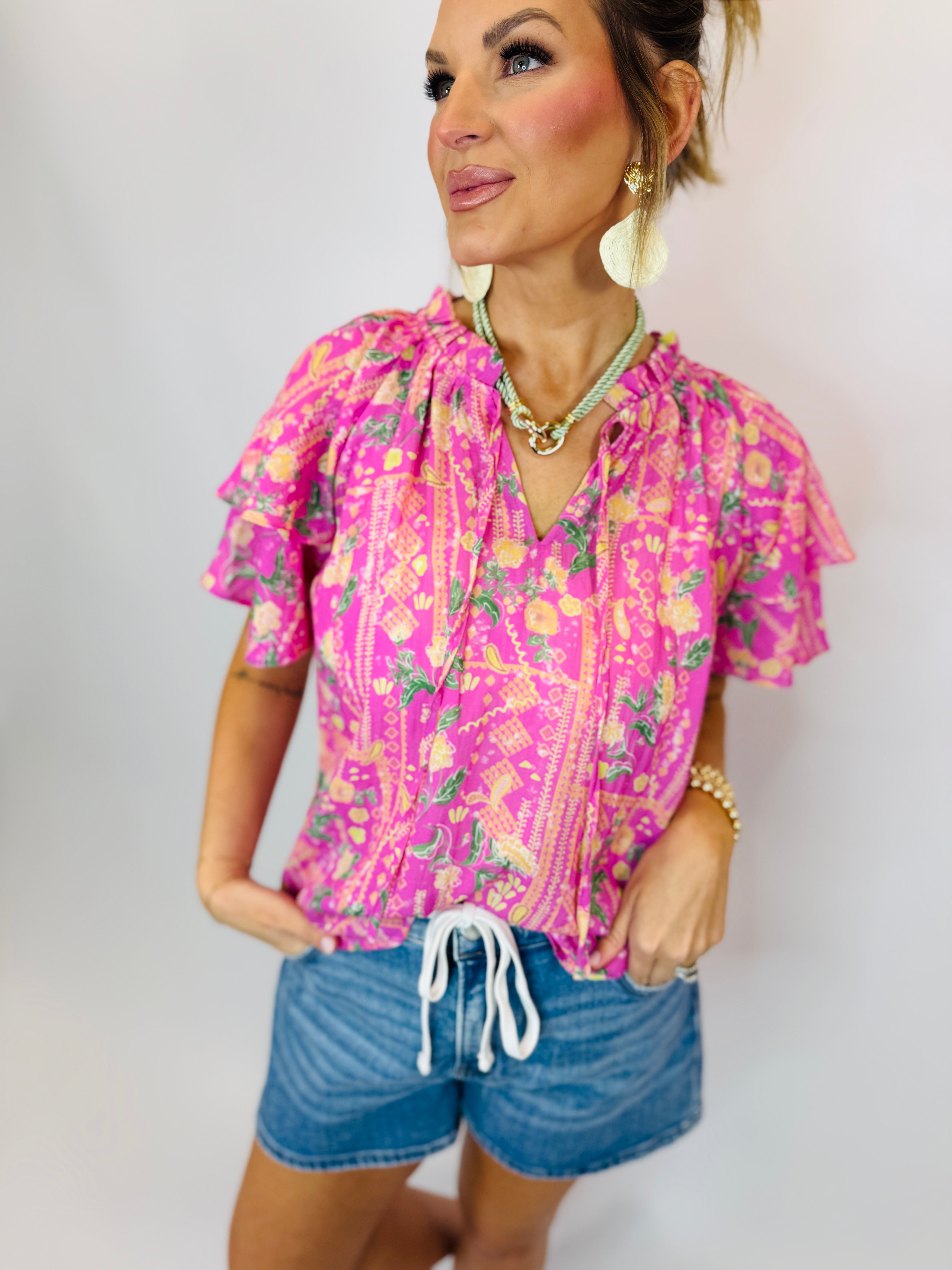 Woman wearing bright pink floral Denisse top with ruffled sleeves and drawstring hem, paired with denim shorts