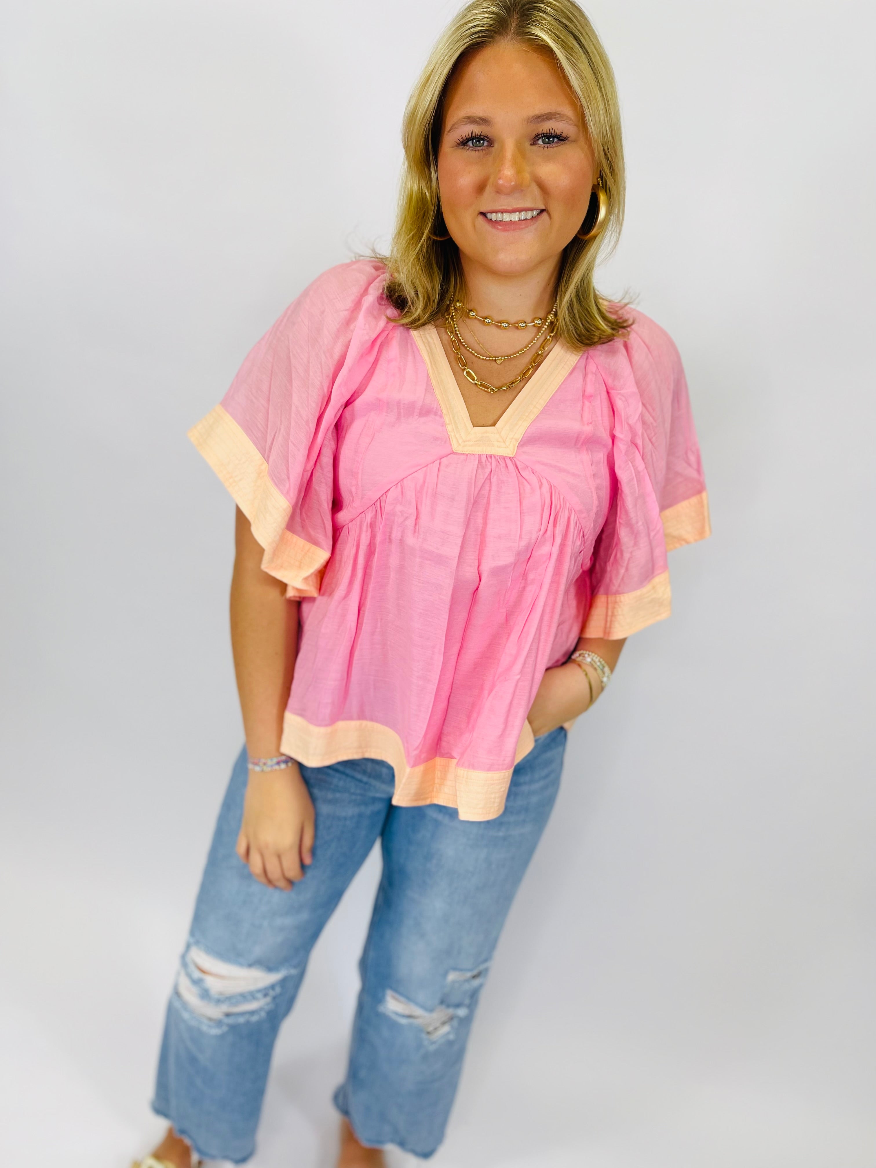 Woman wearing pink flutter sleeve top with peach trim and distressed blue jeans, styled with layered gold necklaces.