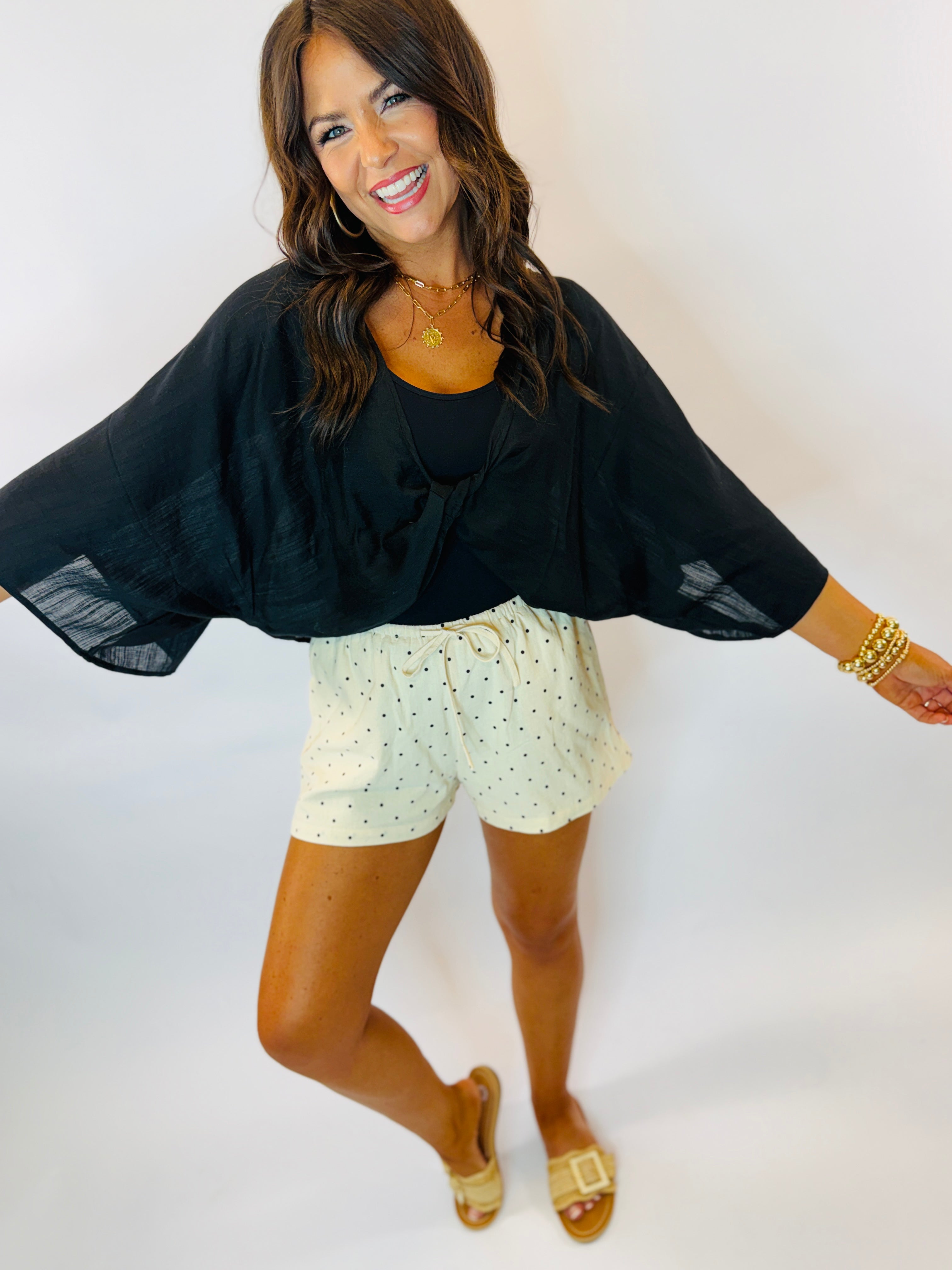 Woman wearing black Harper tie top with flowing sleeves paired with white polka dot shorts and gold sandals