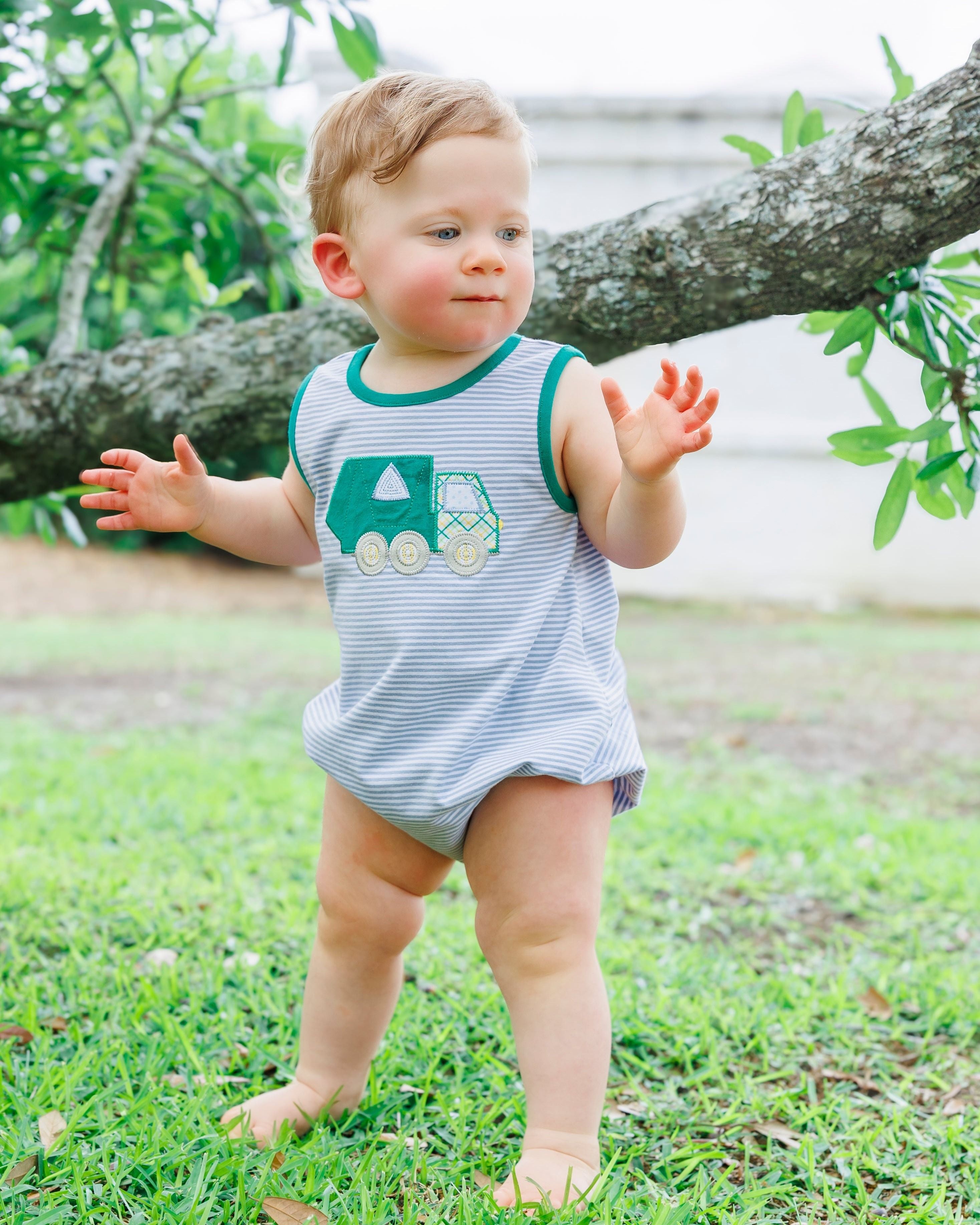 Toddler wearing striped bubble romper with green garbage truck appliqué standing outdoors on grass