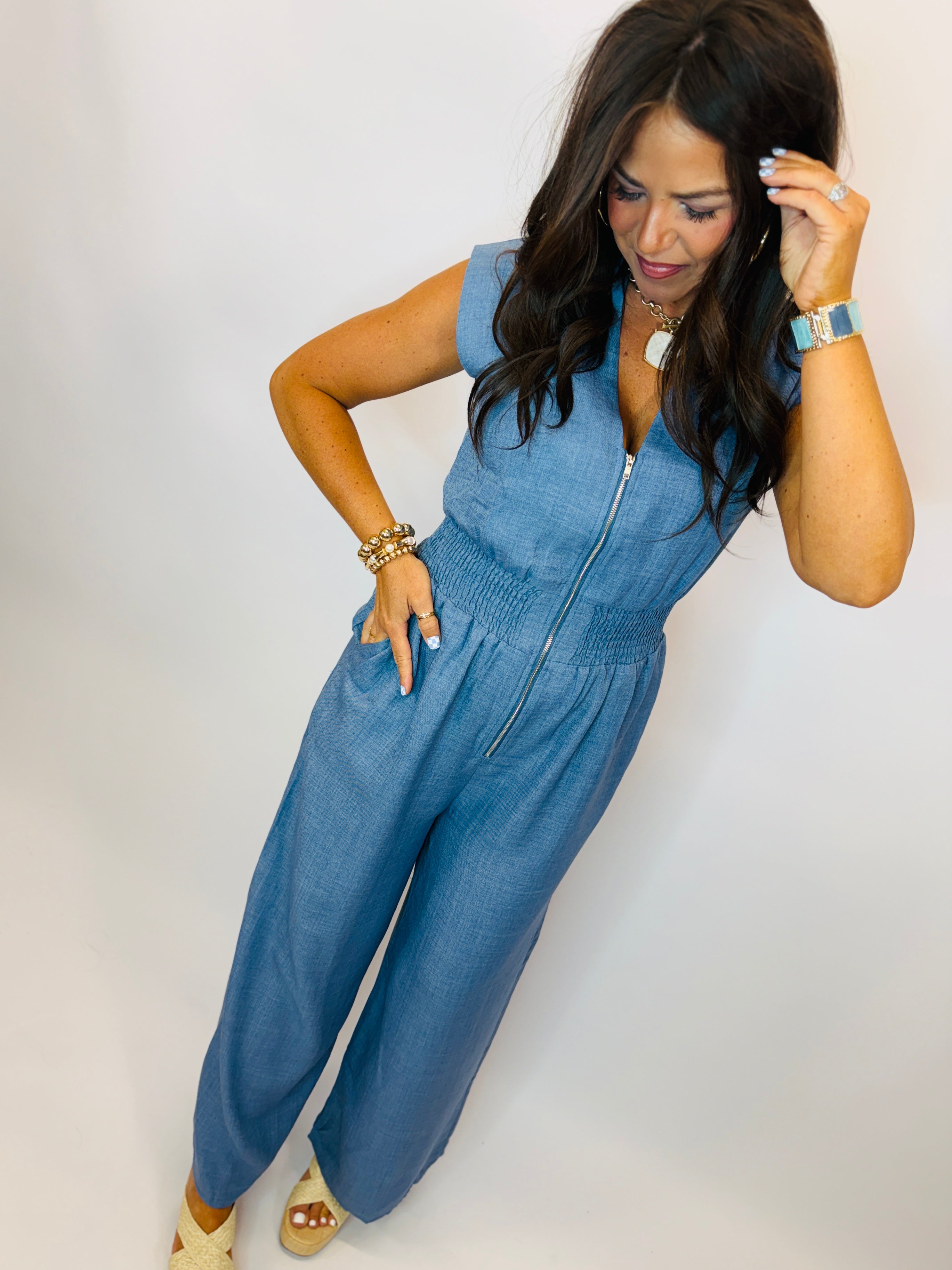 Woman wearing blue denim sleeveless jumpsuit with zip front and smocked waist, styled with gold jewelry and wedge sandals.