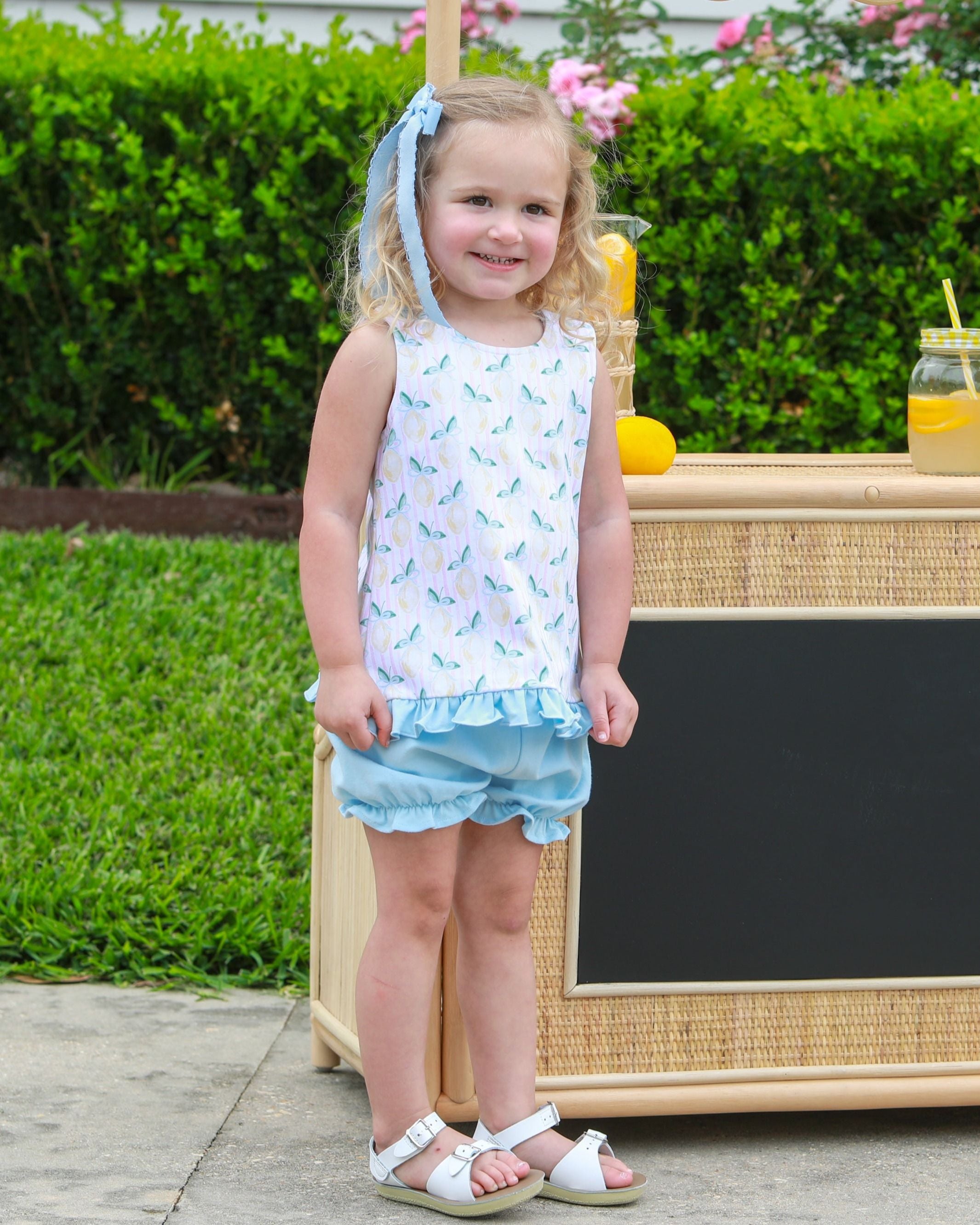 Young girl wearing white tank top with blue pattern and matching blue ruffled bloomers, standing by lemonade stand.