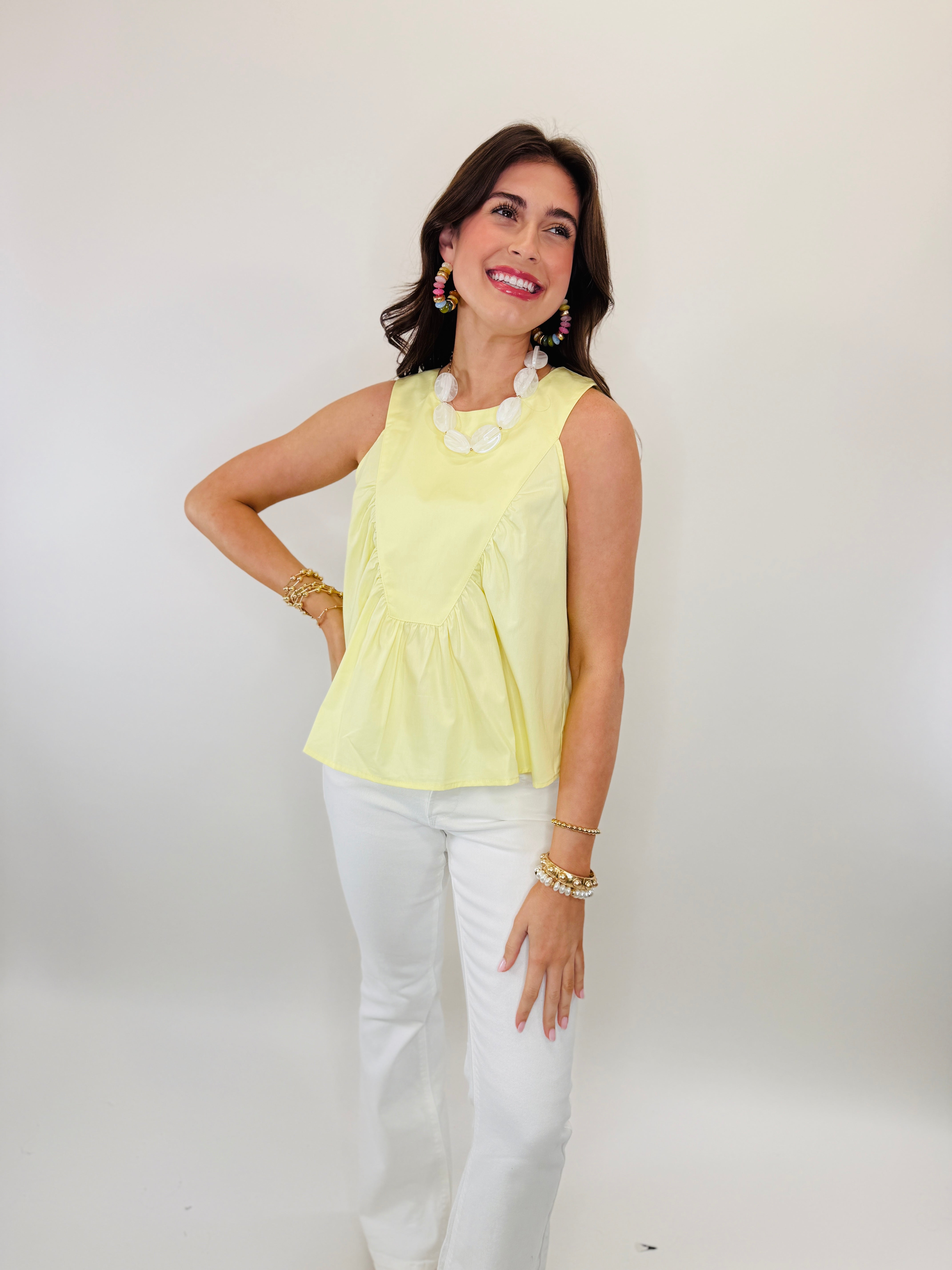 Woman wearing yellow sleeveless Fran top with white pants, pearl necklace, and colorful statement earrings.