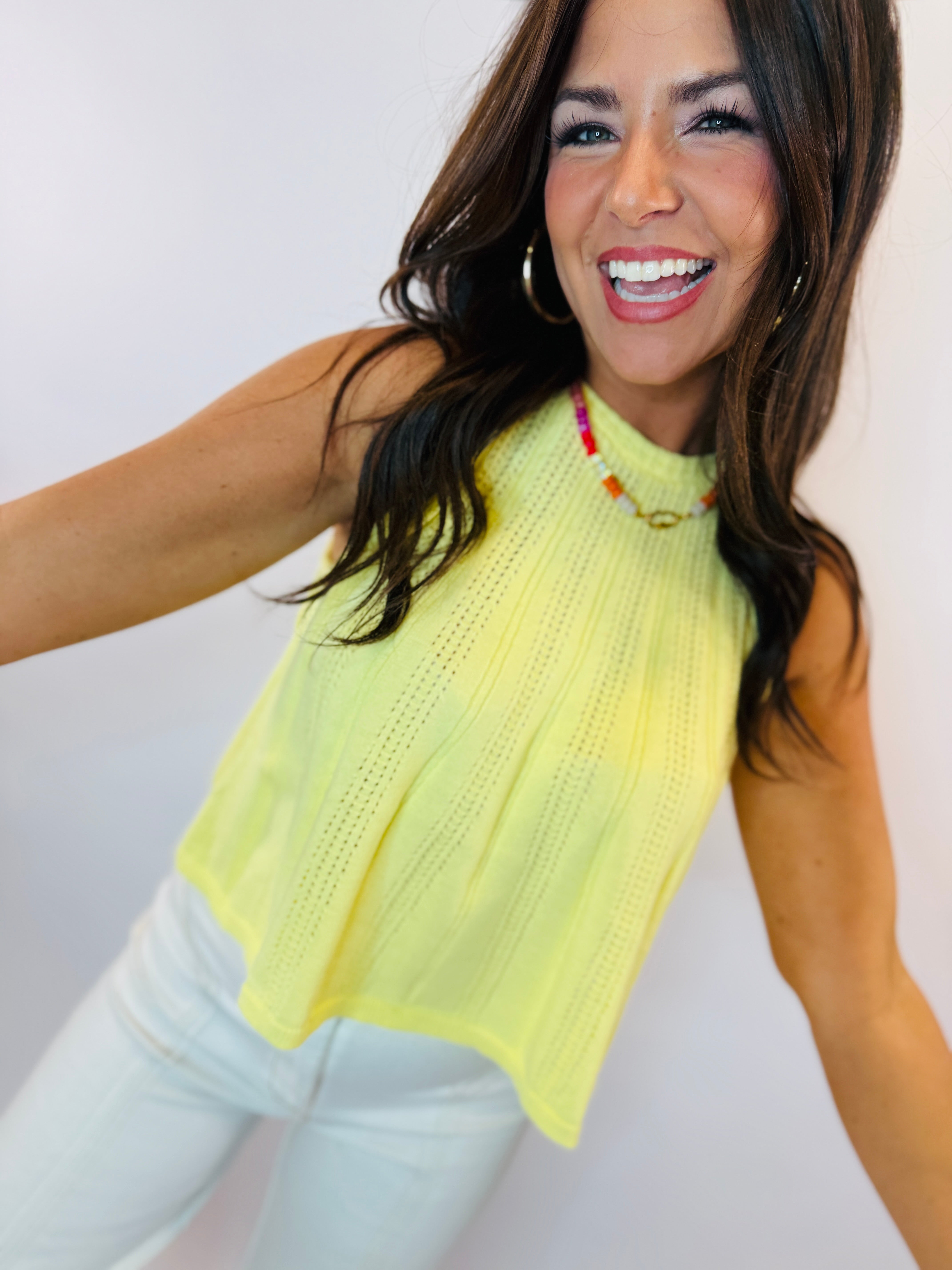 Woman wearing bright yellow knitted sleeveless Grace top with white pants, smiling against white background.