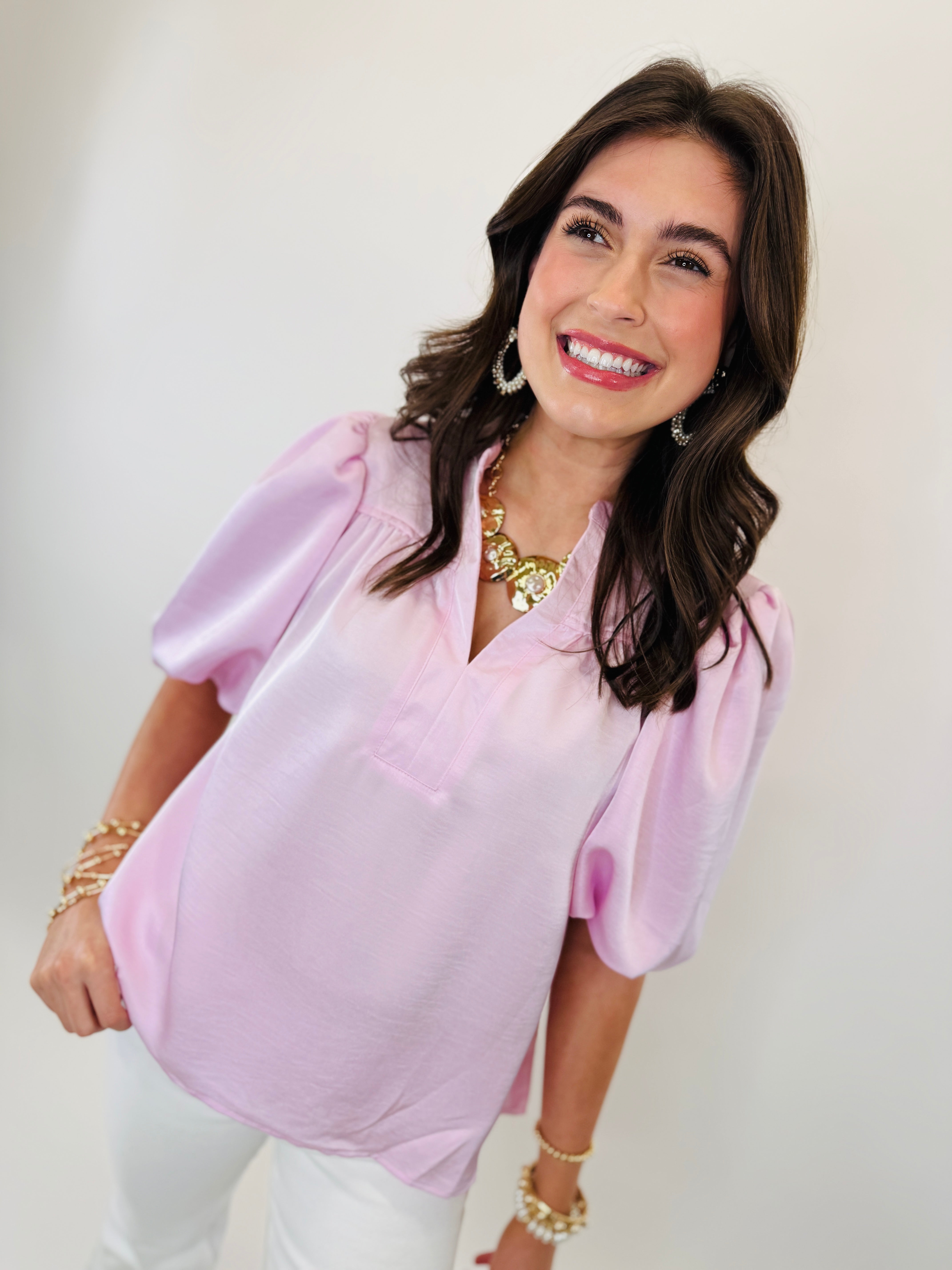 Woman wearing pink puffy sleeve blouse with gold jewelry and pearl earrings, smiling at camera