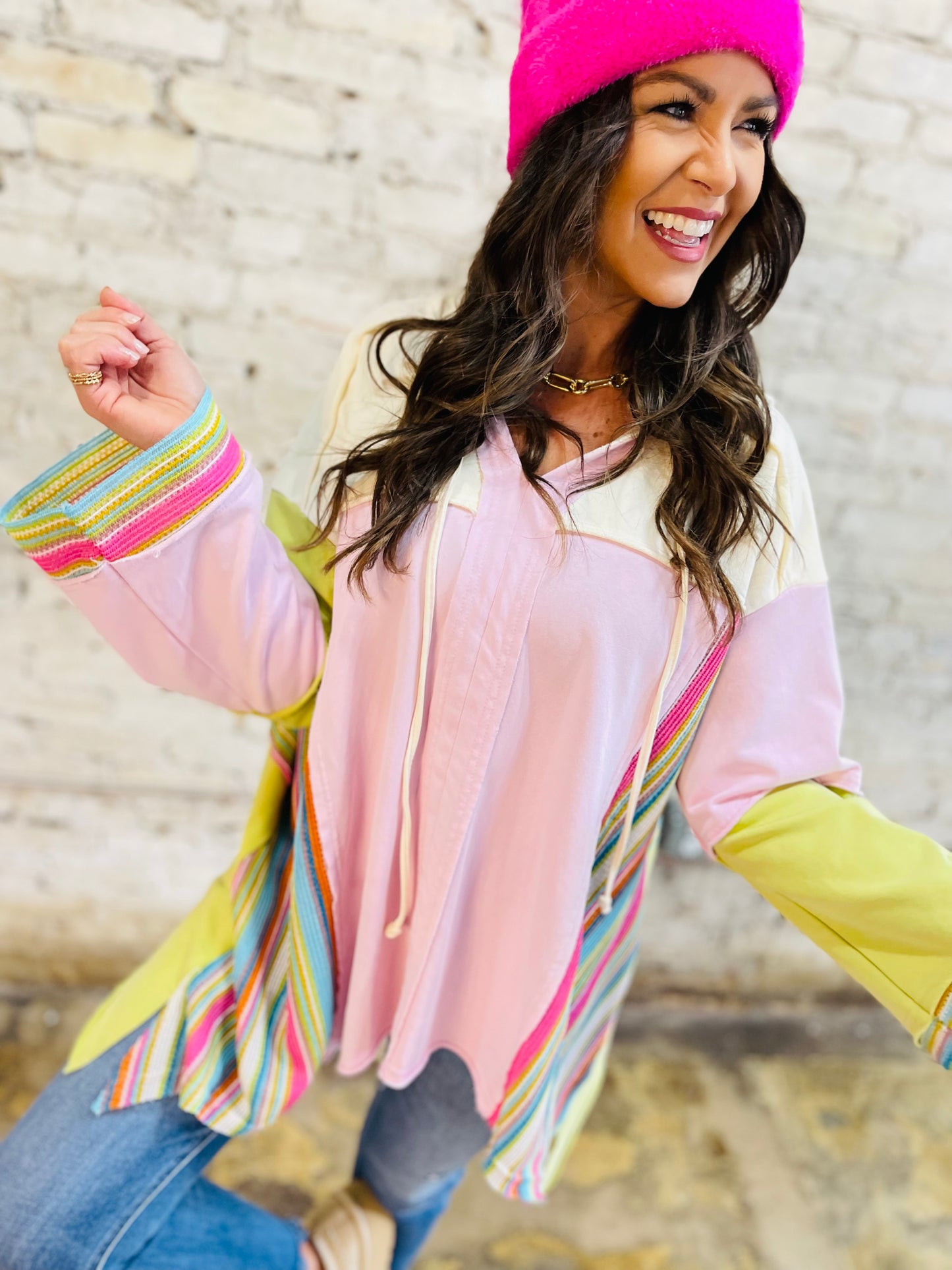 Woman wearing colorful striped Ember hoodie with pink beanie, smiling and posing against brick wall background.