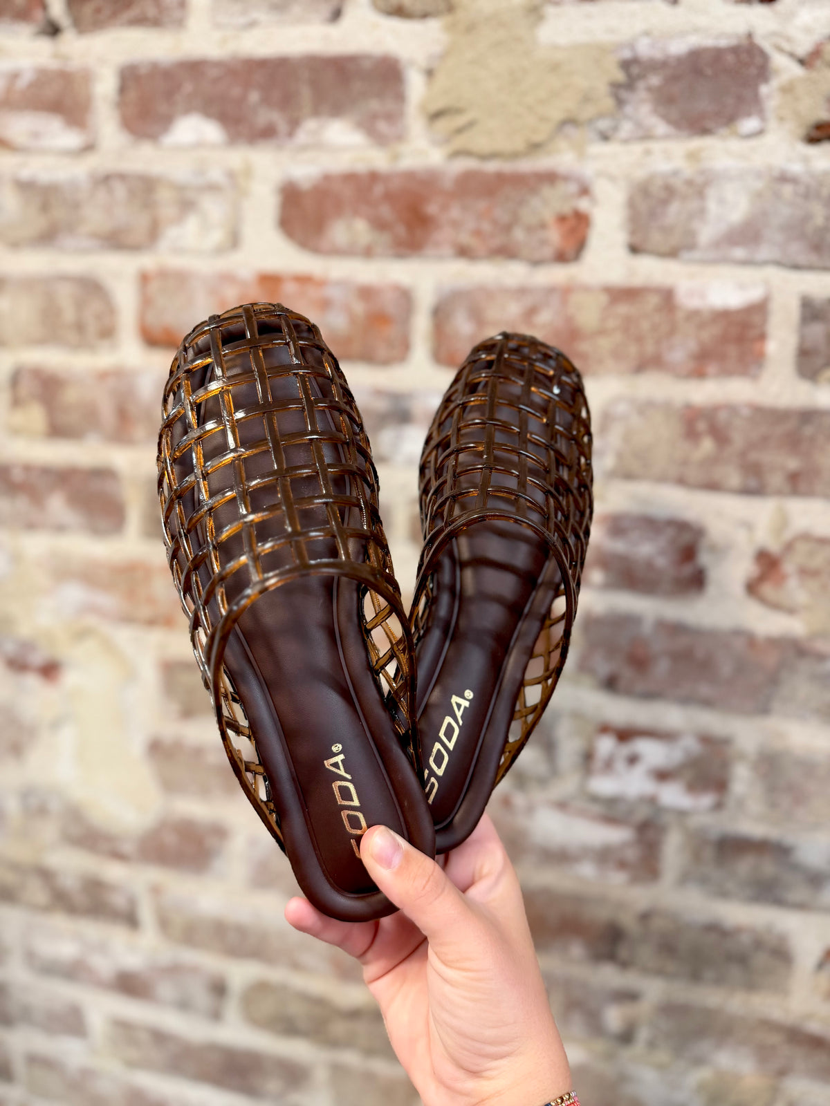 Soda brand Jane Slip On brown translucent caged jelly mule shoes held against brick wall.