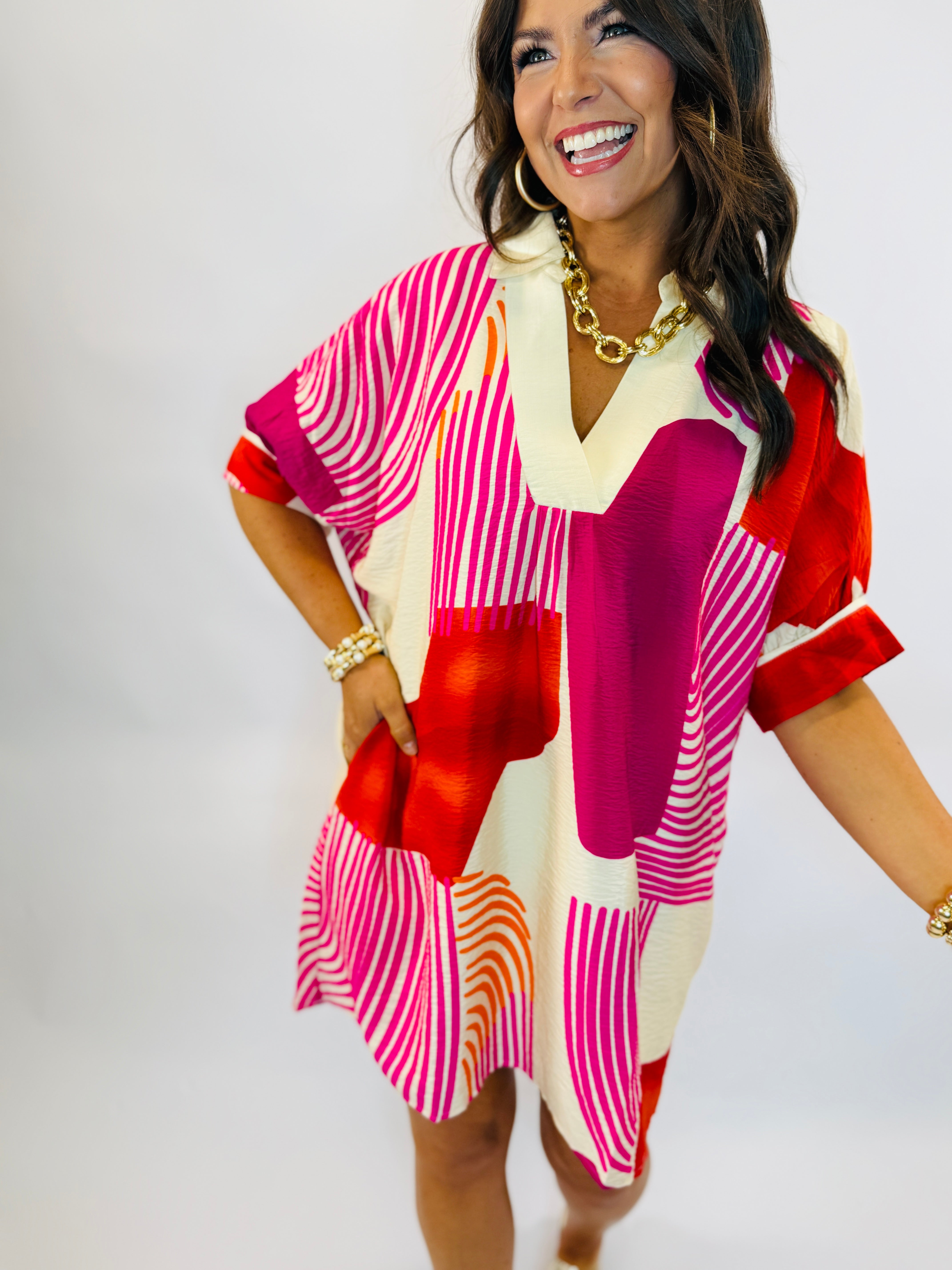 Woman wearing colorful Hannah dress with geometric patterns in pink, red, orange and cream with gold chain necklace.
