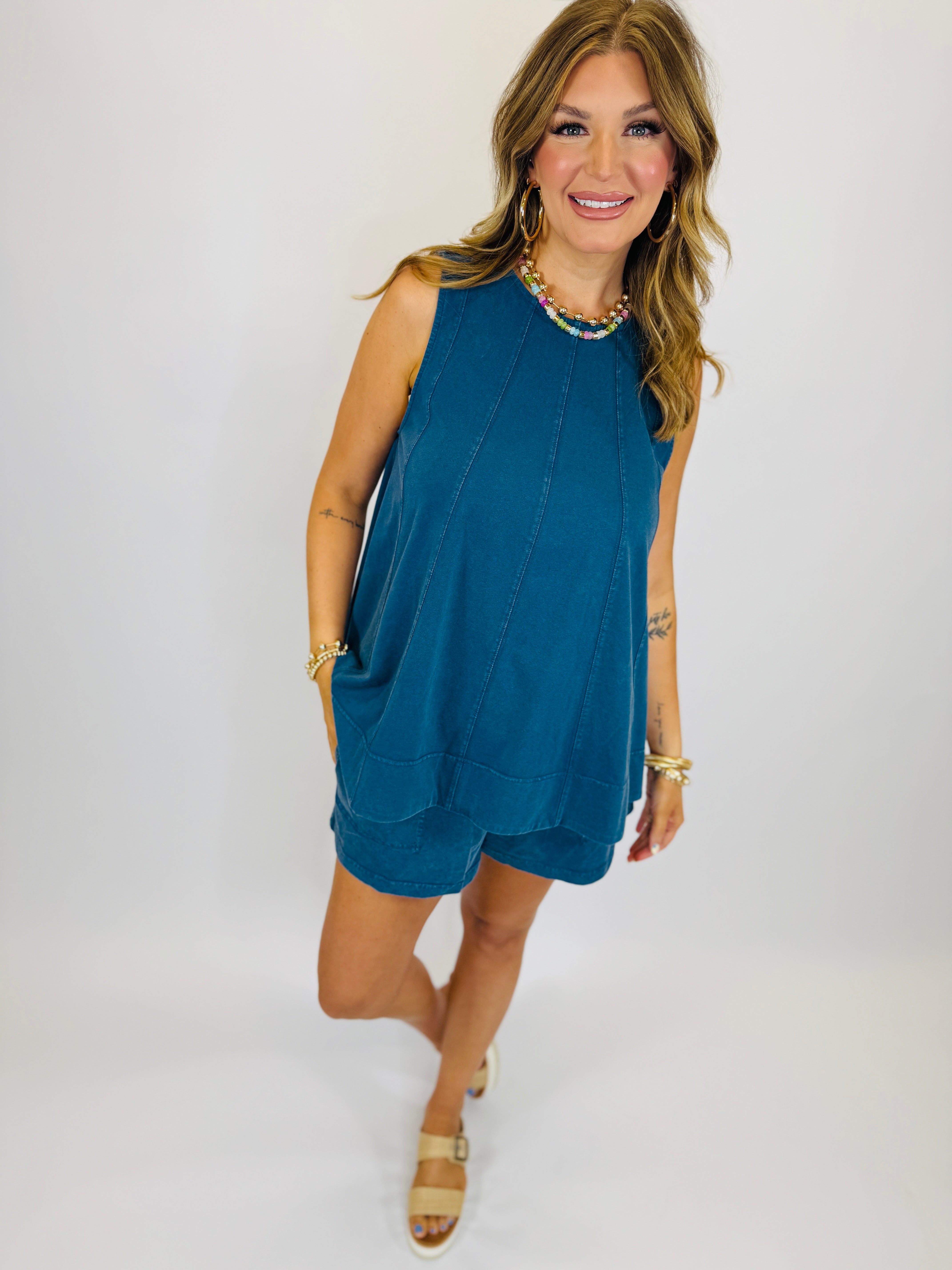 Woman wearing blue sleeveless shift dress with scalloped hem, gold jewelry, and tan strappy sandals