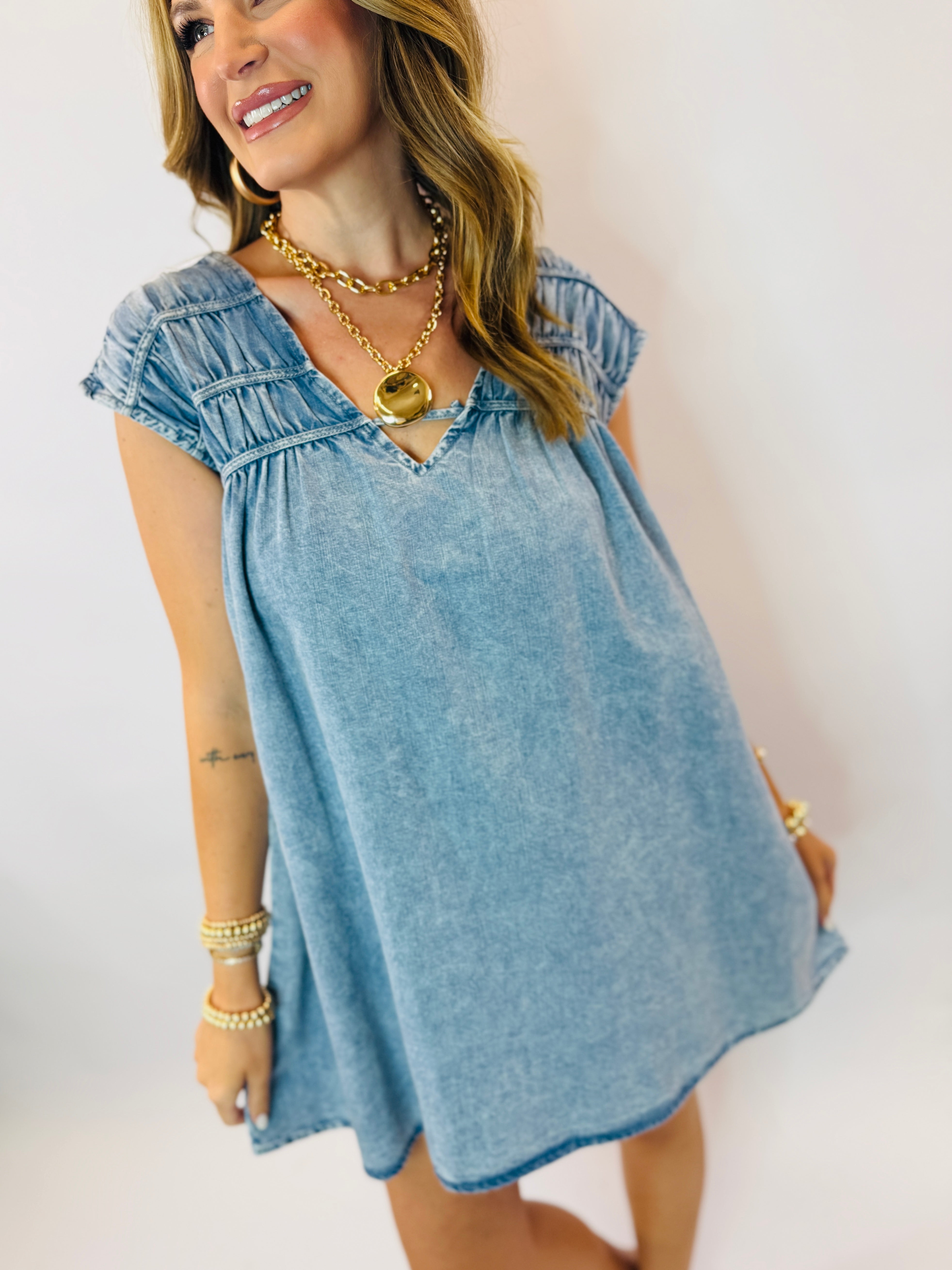 Woman wearing light blue denim dress with cap sleeves, V-neck, and layered gold chain necklaces.