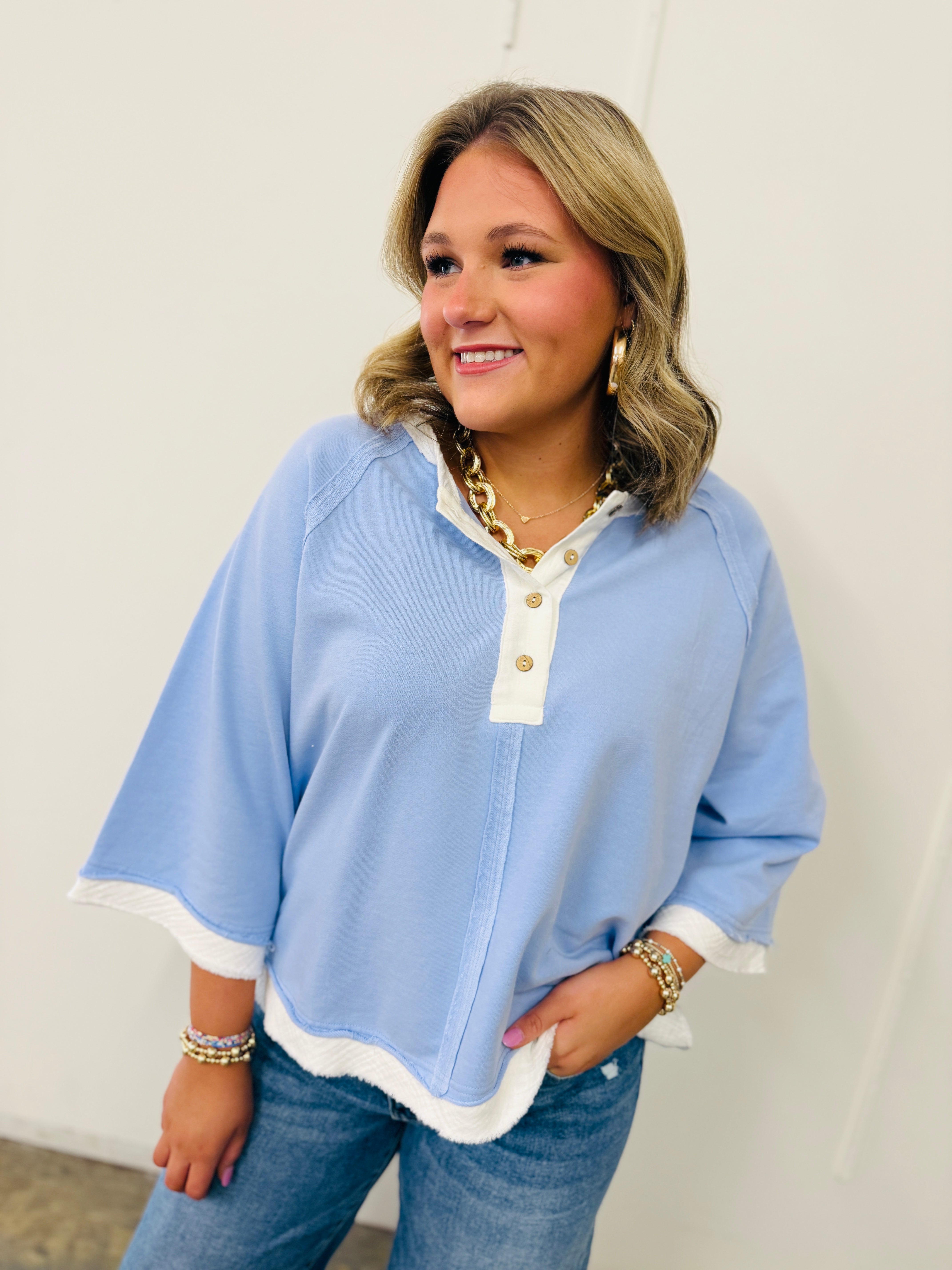 Woman wearing light blue Elizabeth top with white contrast trim and gold button details, paired with jeans and gold jewelry.