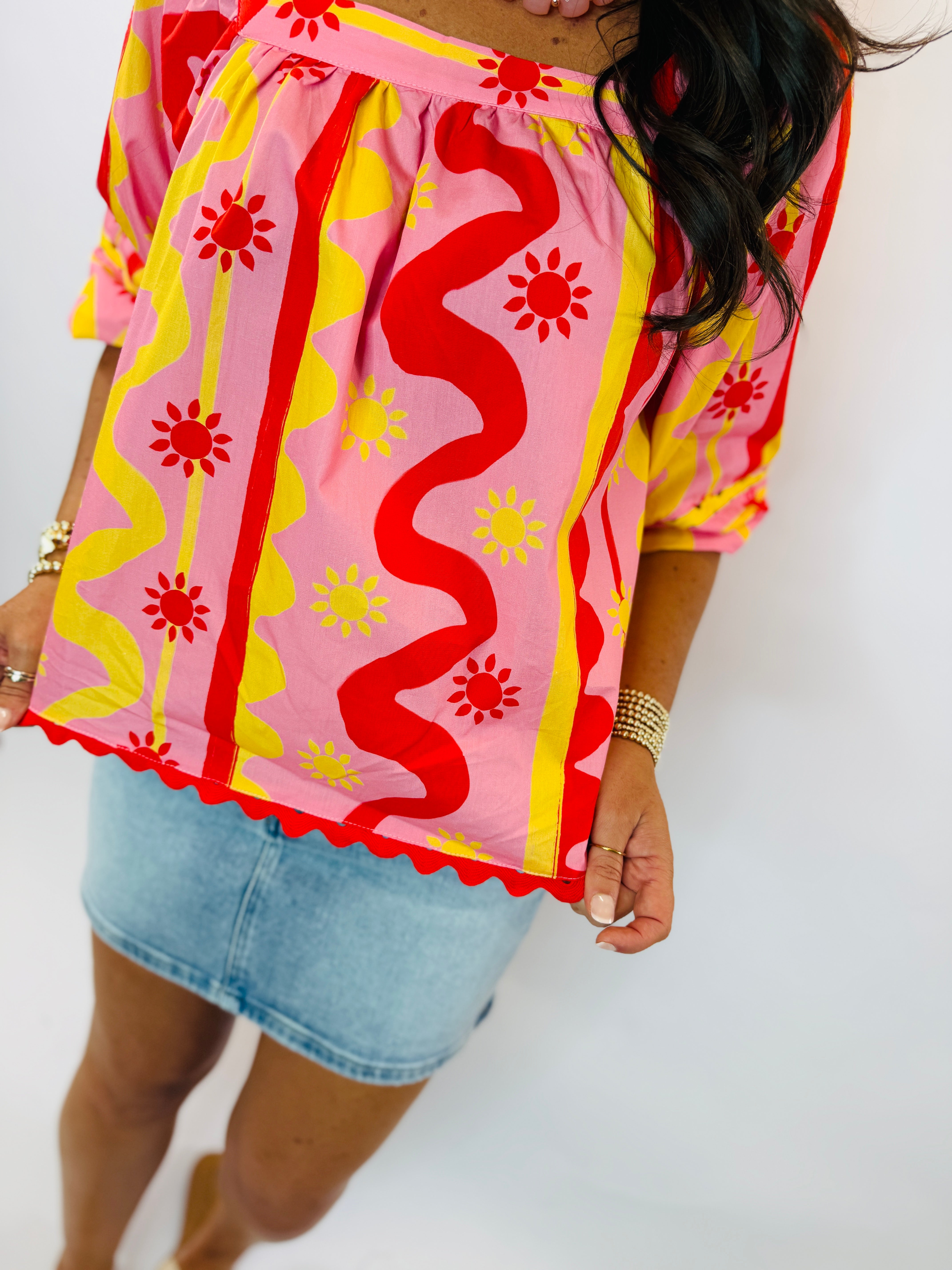 Woman wearing colorful off-shoulder Emmie top with pink, red, and yellow wavy pattern and sun motifs, paired with denim sk...