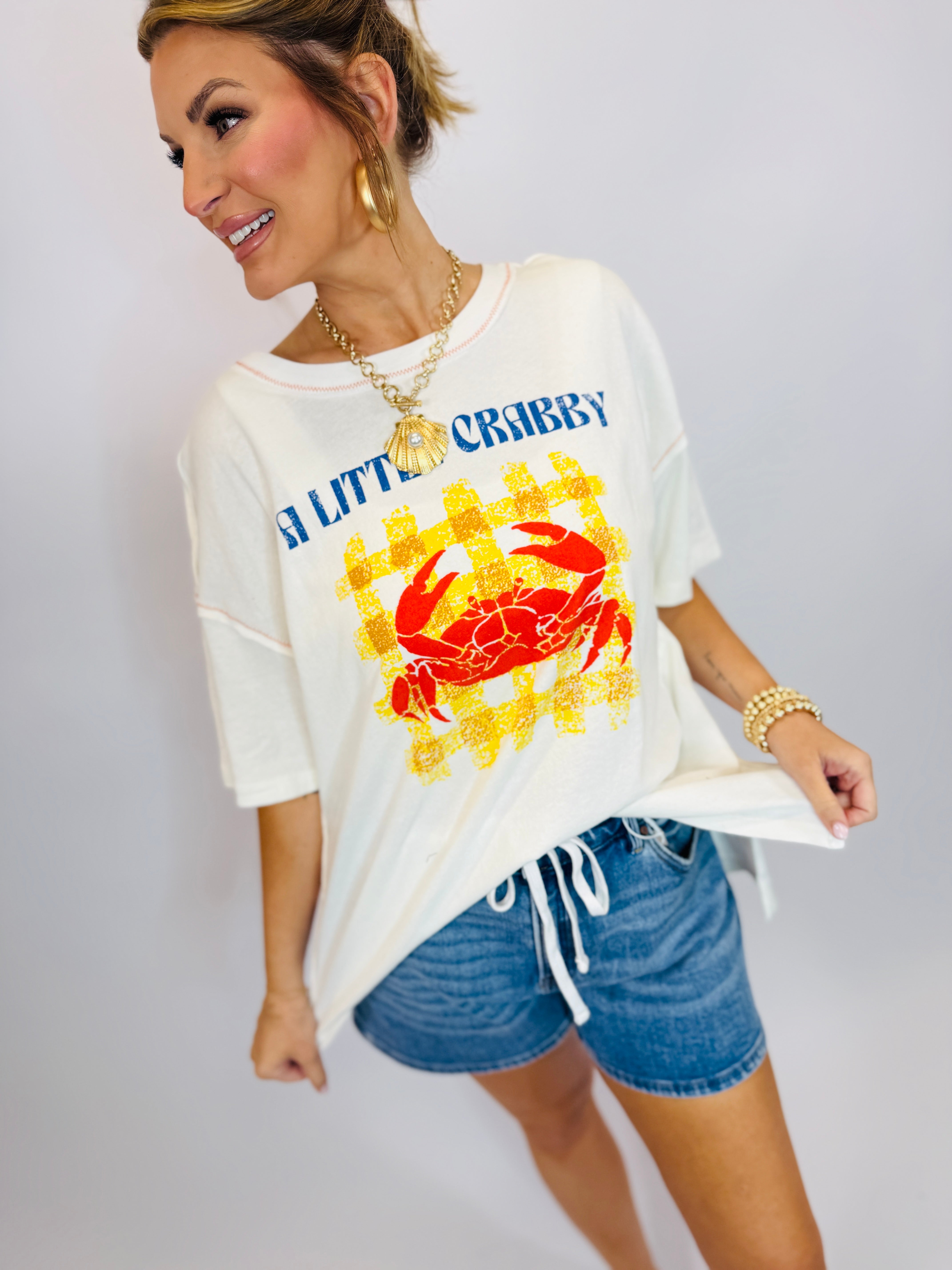 Woman wearing white oversized "A Little Crabby" graphic tee with red crab design and blue denim shorts.