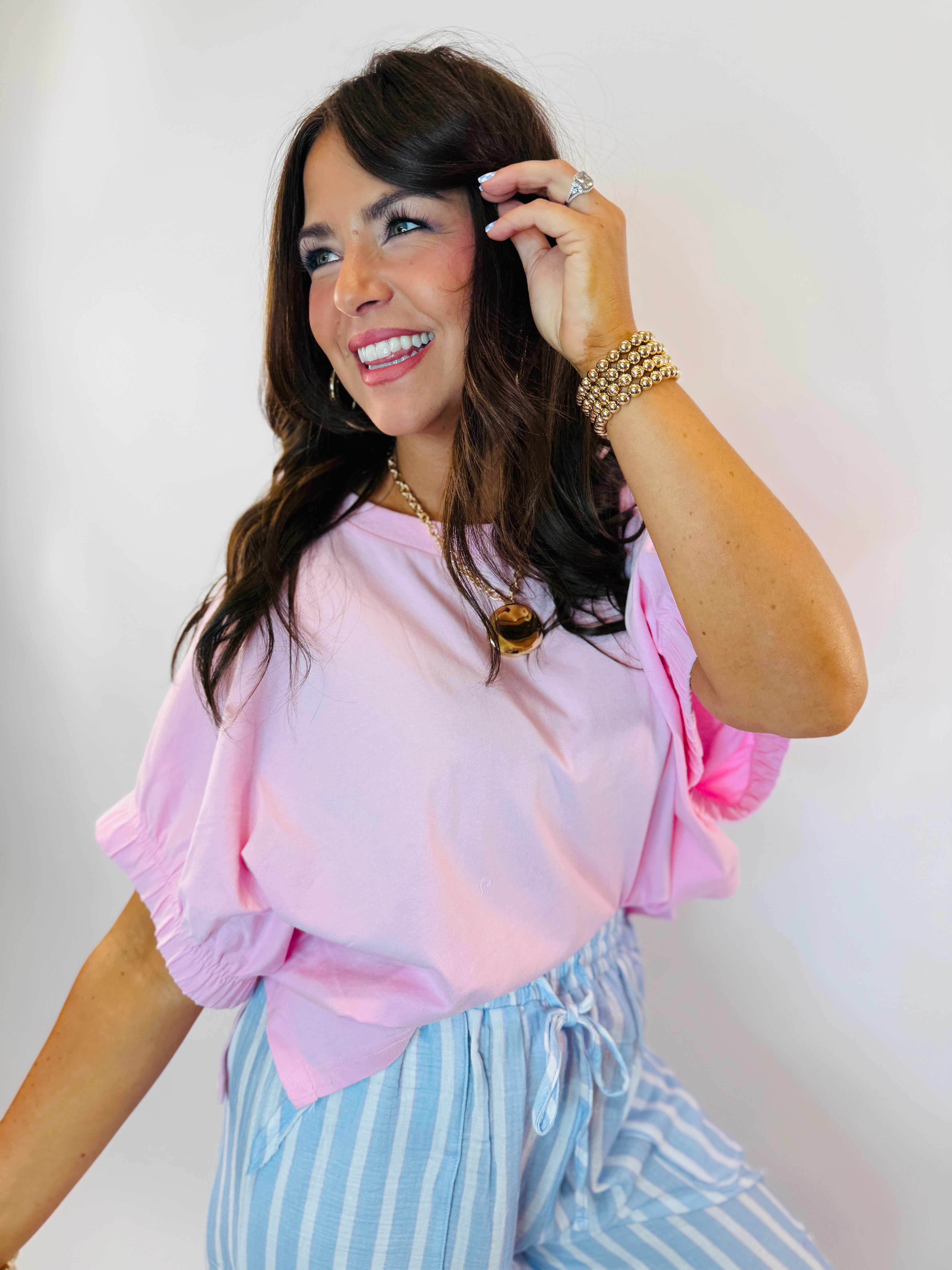 Woman wearing pink Lucy top with ruffled sleeves, styled with blue striped bottoms and gold jewelry accessories.