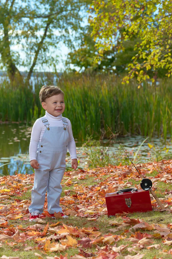 Young child wearing light blue striped overalls and white shirt standing by autumn lake with fishing gear.