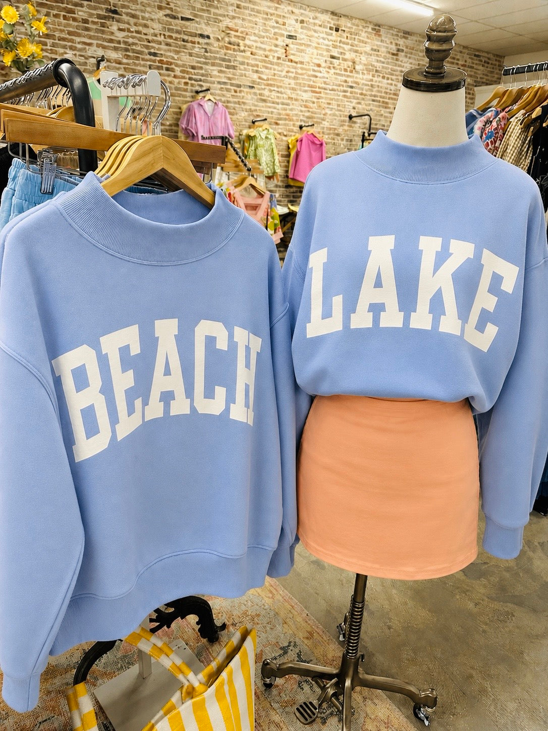 Light blue mock neck sweatshirts with "BEACH" and "LAKE" text displayed on hangers and mannequin in clothing store